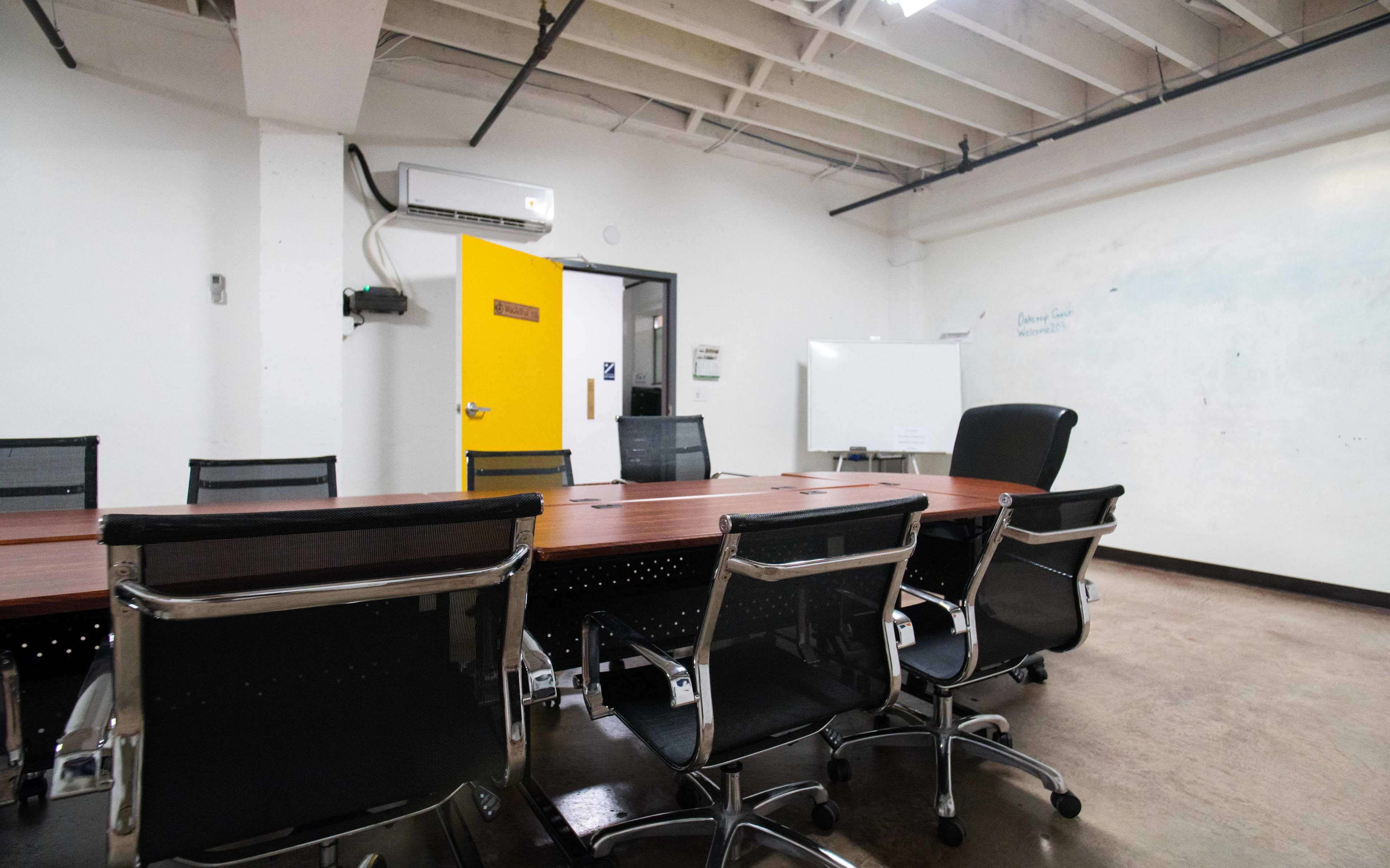 A conference room features a long wooden table surrounded by black office chairs, with a yellow door and a whiteboard in the background.
