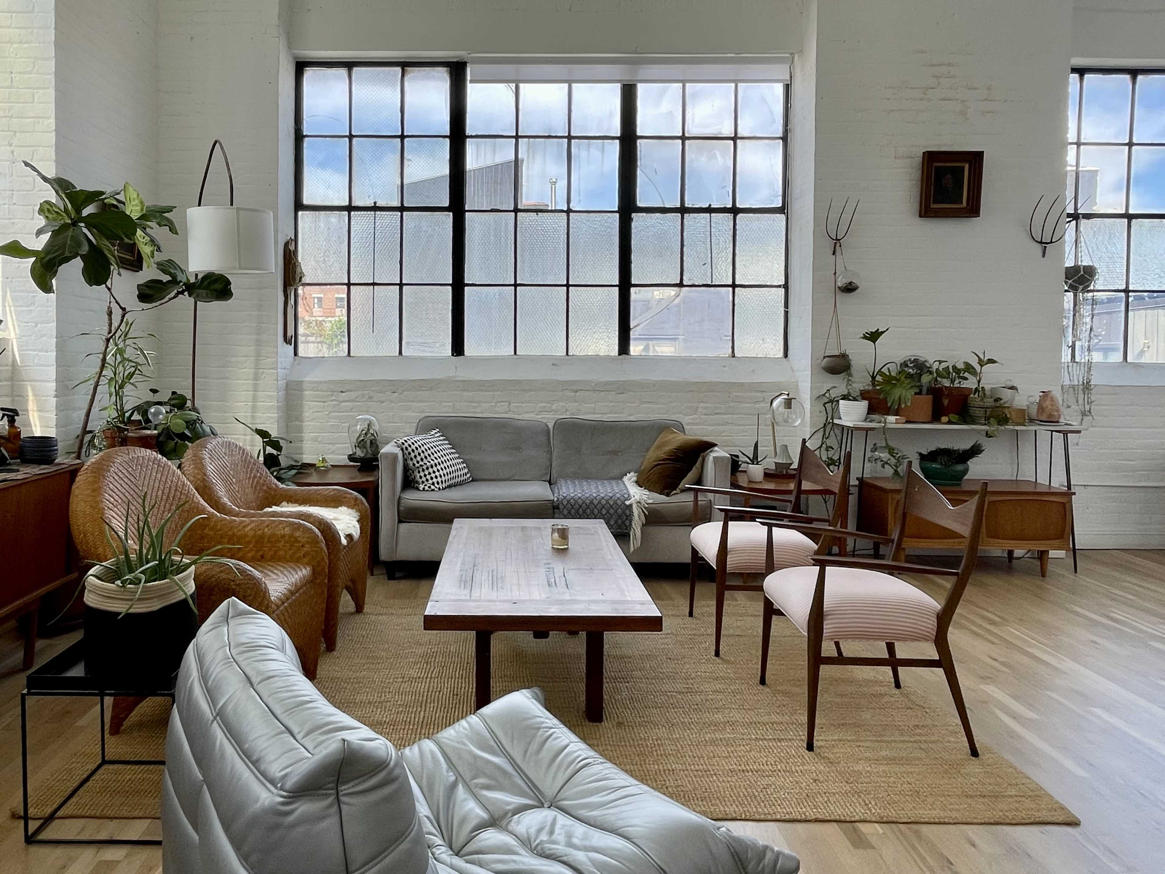 The image shows a spacious, well-lit living room featuring a mix of modern and vintage furniture, large windows, and various plants.
