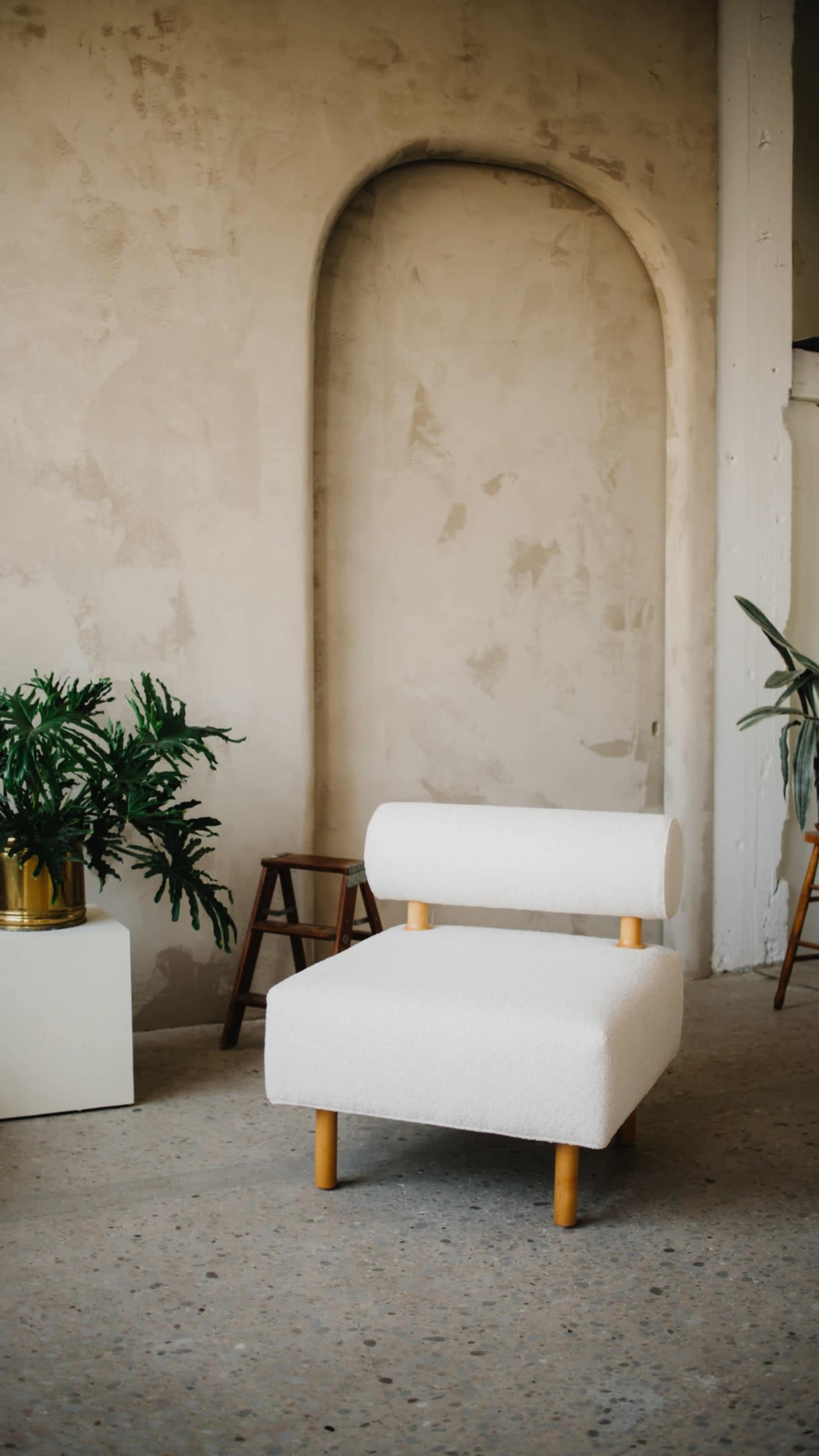 A minimalist room with a white upholstered chair, a small wooden stool, and two potted plants against a textured wall.