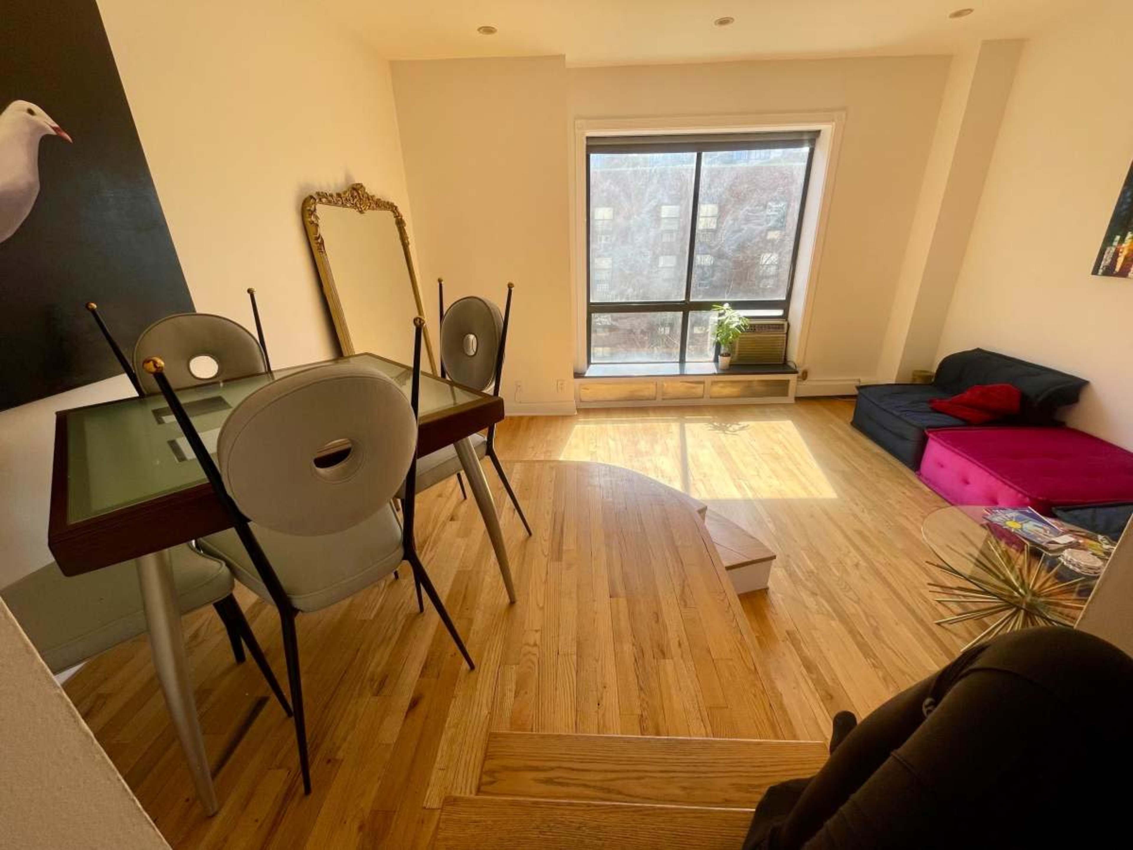 The image shows a bright, modern room featuring a glass dining table with four chairs, a large window, a dark sofa, and wooden flooring.