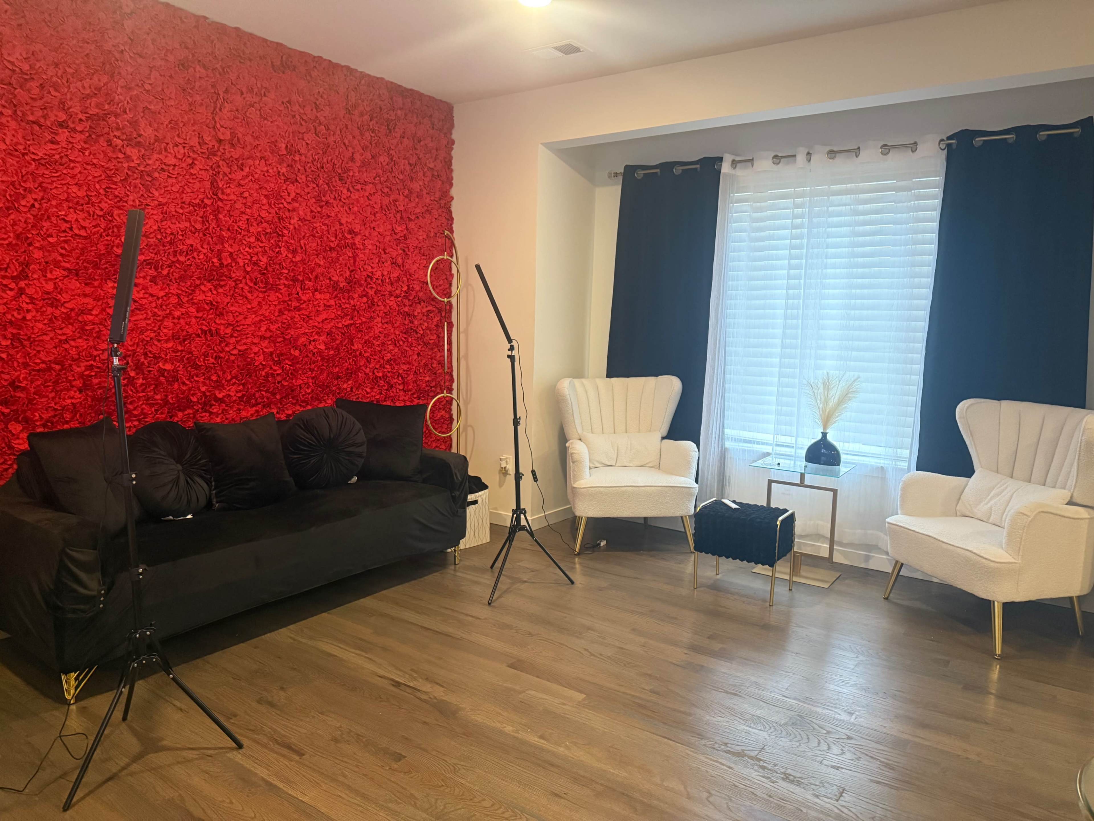 The room features a black couch against a red floral accent wall, two white armchairs, and a small table beside a window with sheer curtains.