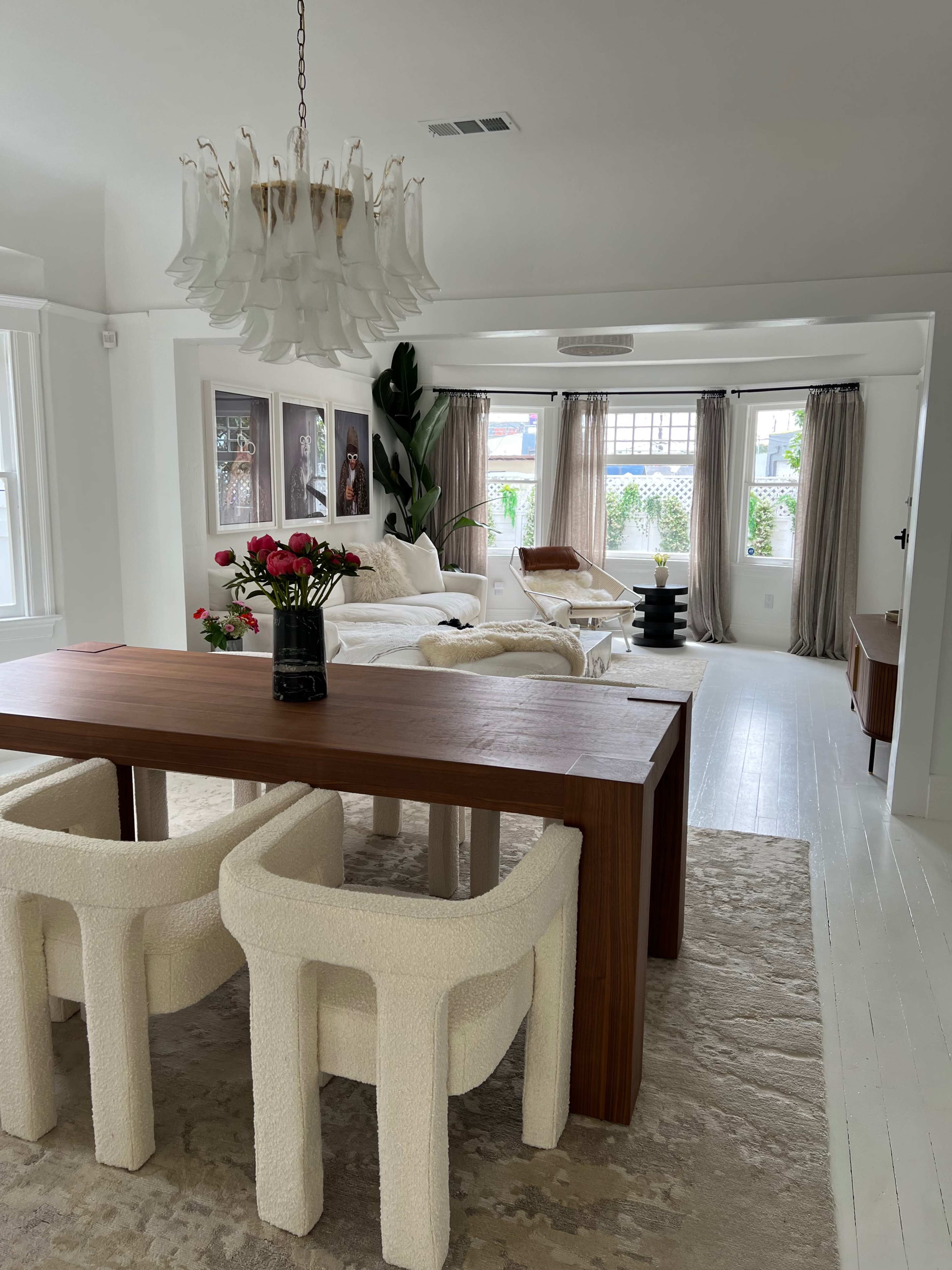 A modern living space featuring a wooden dining table with upholstered chairs, a plush seating area with a light-colored couch, and large windows draped with sheer curtains.