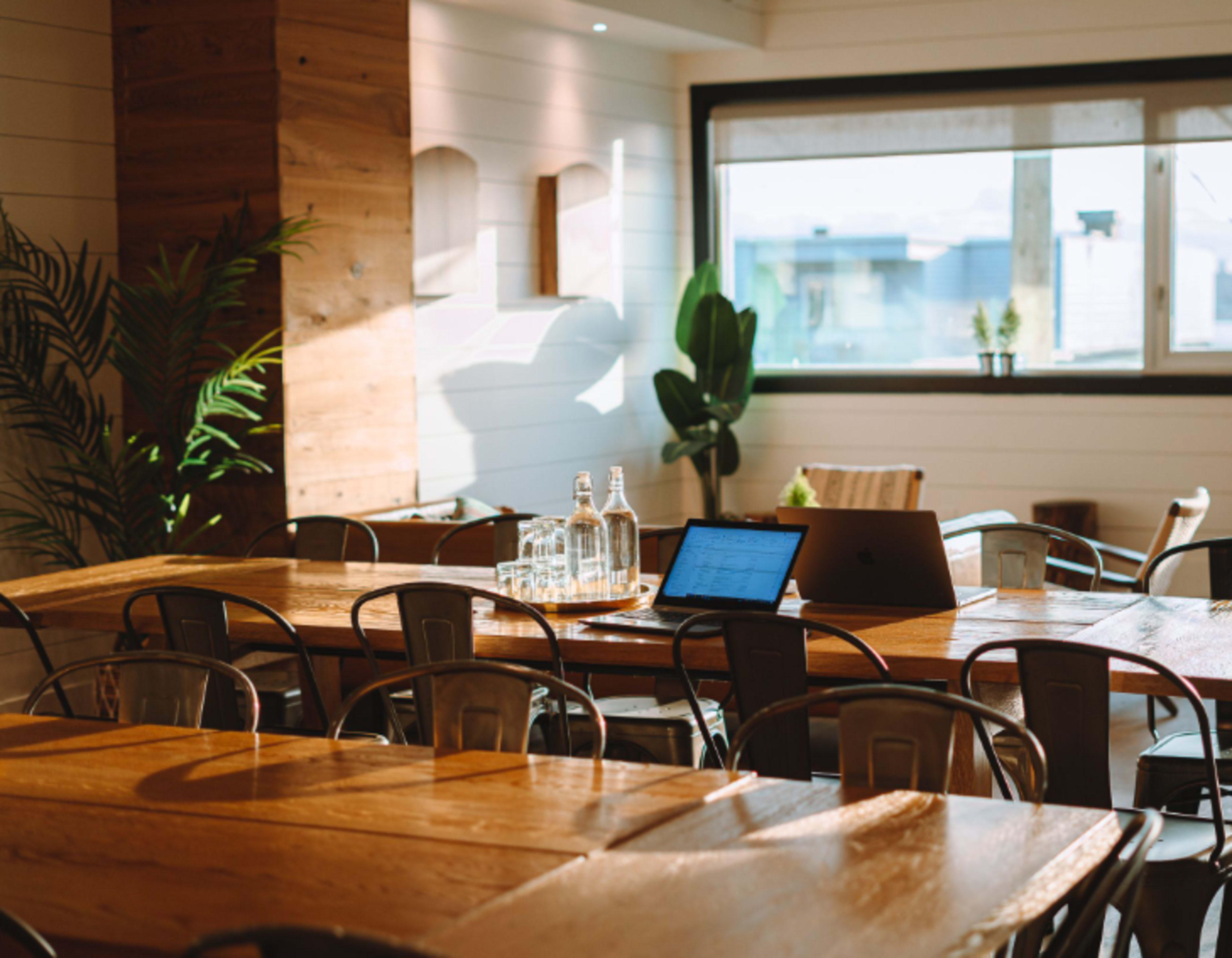 A spacious, well-lit meeting room features wooden tables, metal chairs, and a laptop on a table alongside glass water bottles.