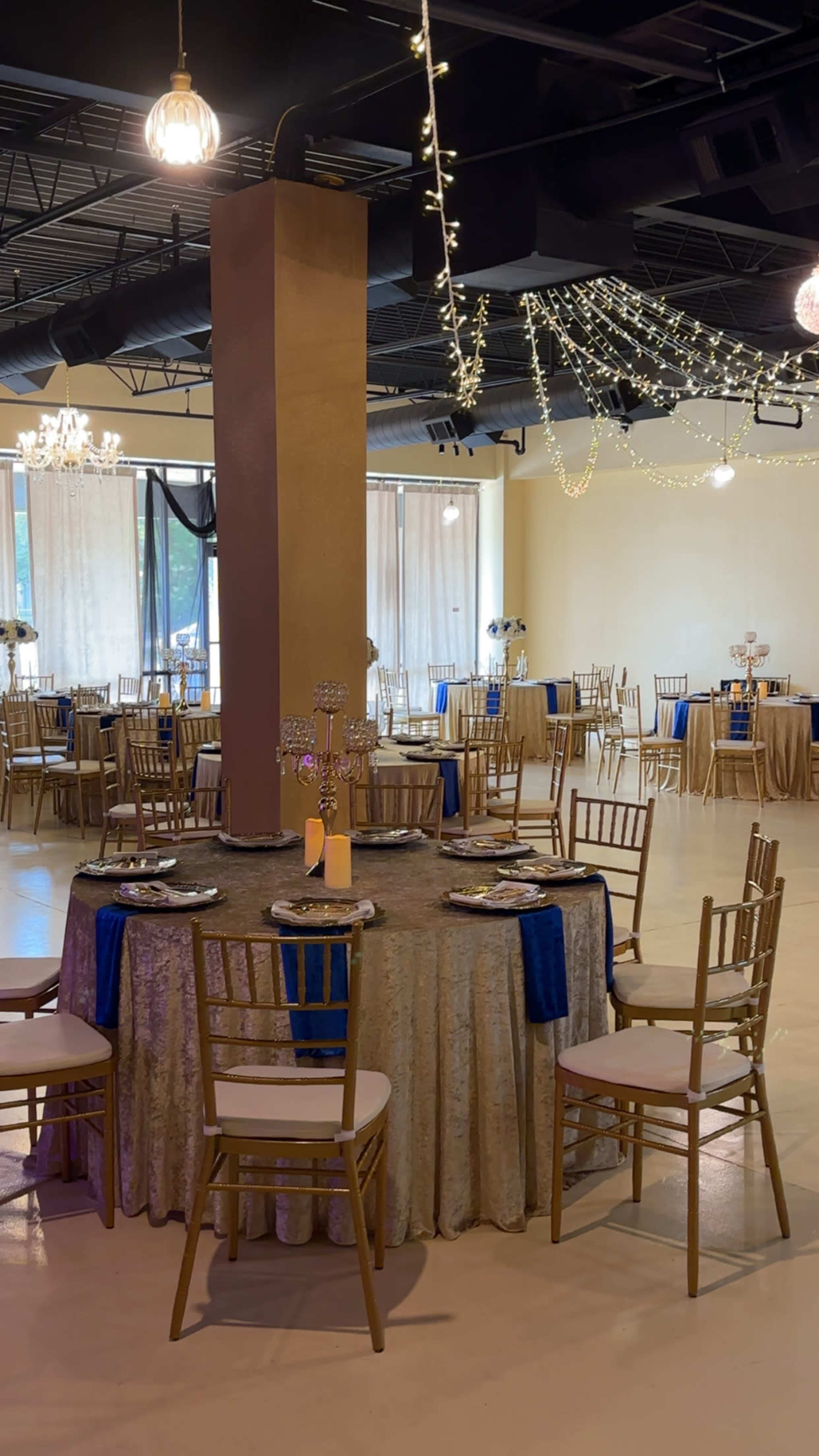 A spacious banquet hall features elegantly set tables with gold and blue decorations, surrounded by chairs and adorned with string lights overhead.