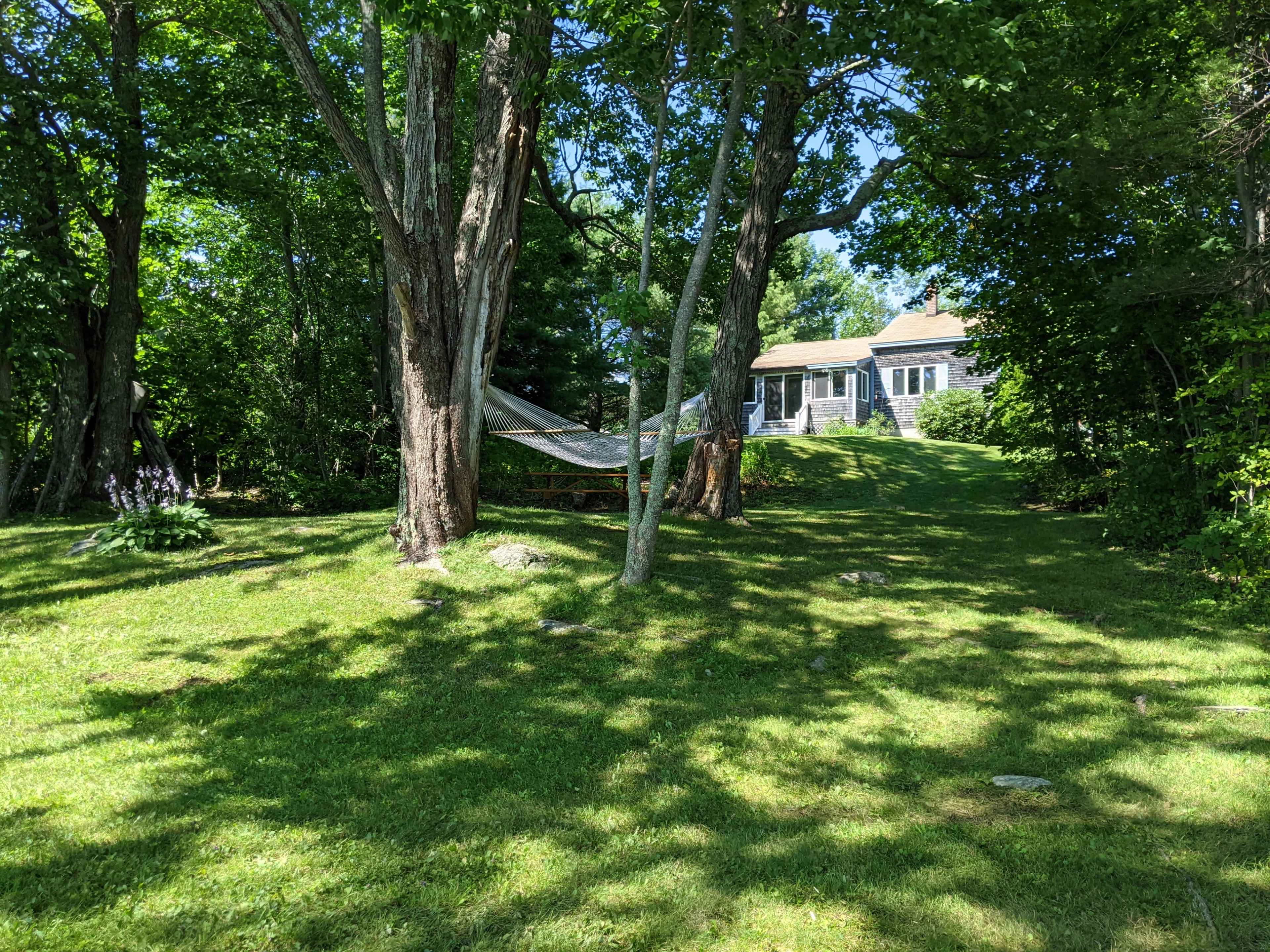 A house with a porch is situated on a grassy area surrounded by large trees and a hammock.