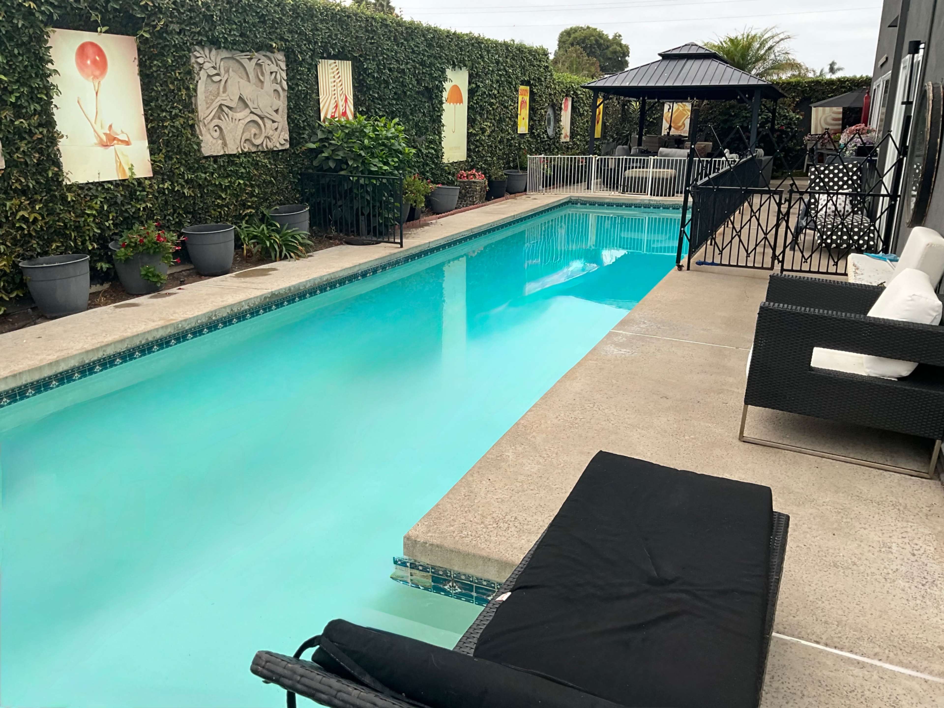 The image shows a swimming pool bordered by potted plants and decorative art on the walls, with a shaded seating area nearby.