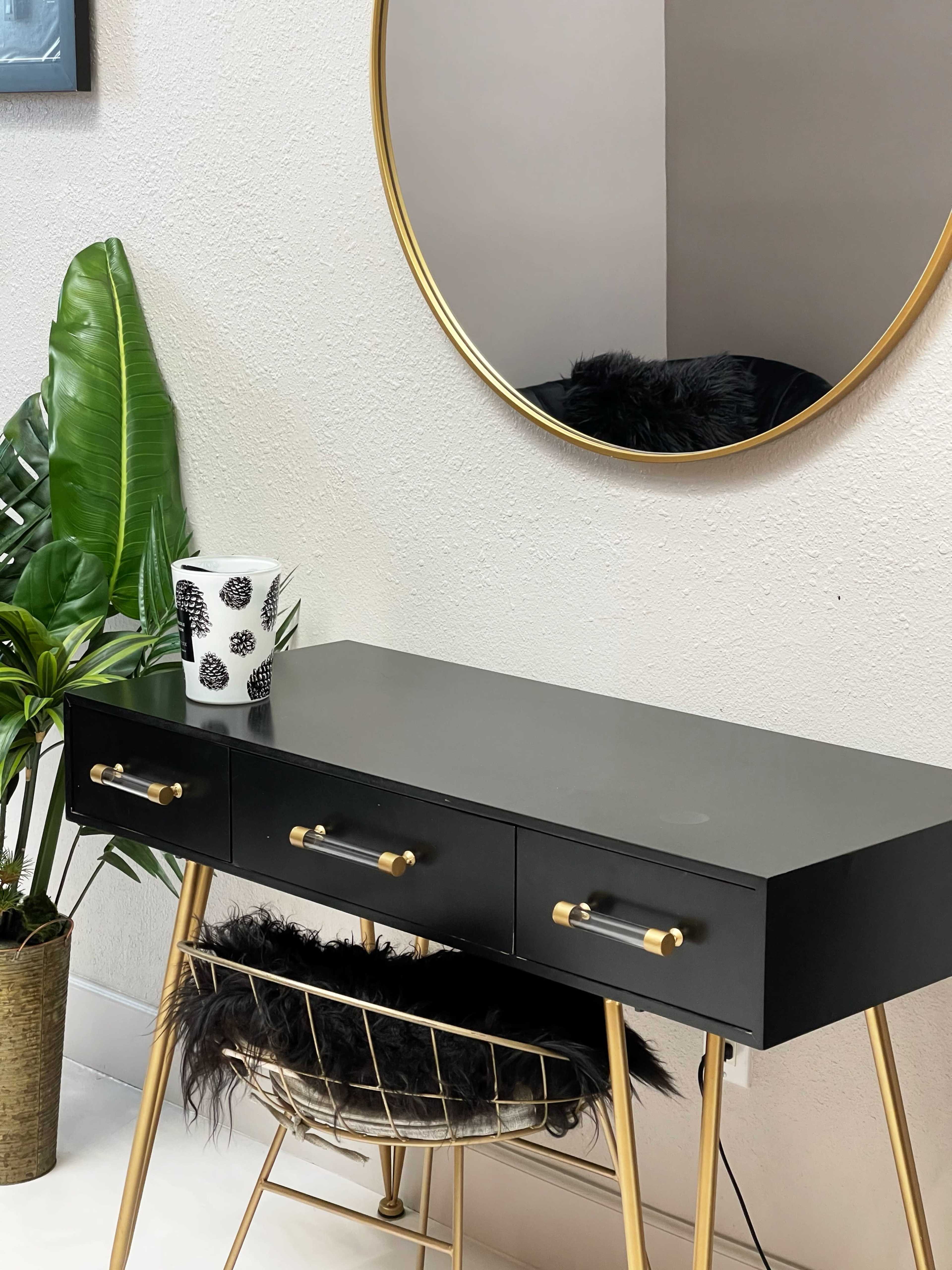 The image features a black vanity table with two drawers, a gold framed round mirror above it, and a potted plant beside a decorative cup on the table.