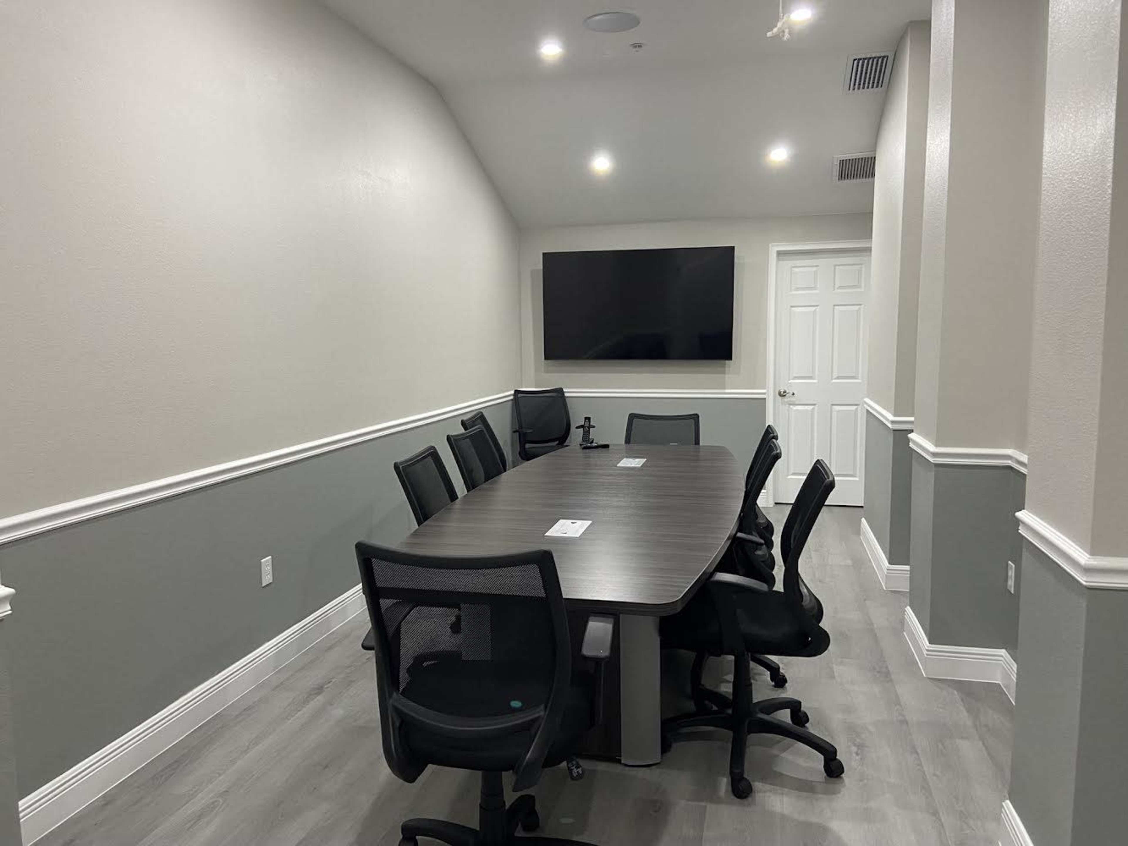 A modern conference room with a long table surrounded by eight office chairs, a wall-mounted screen, and a door at the far end.