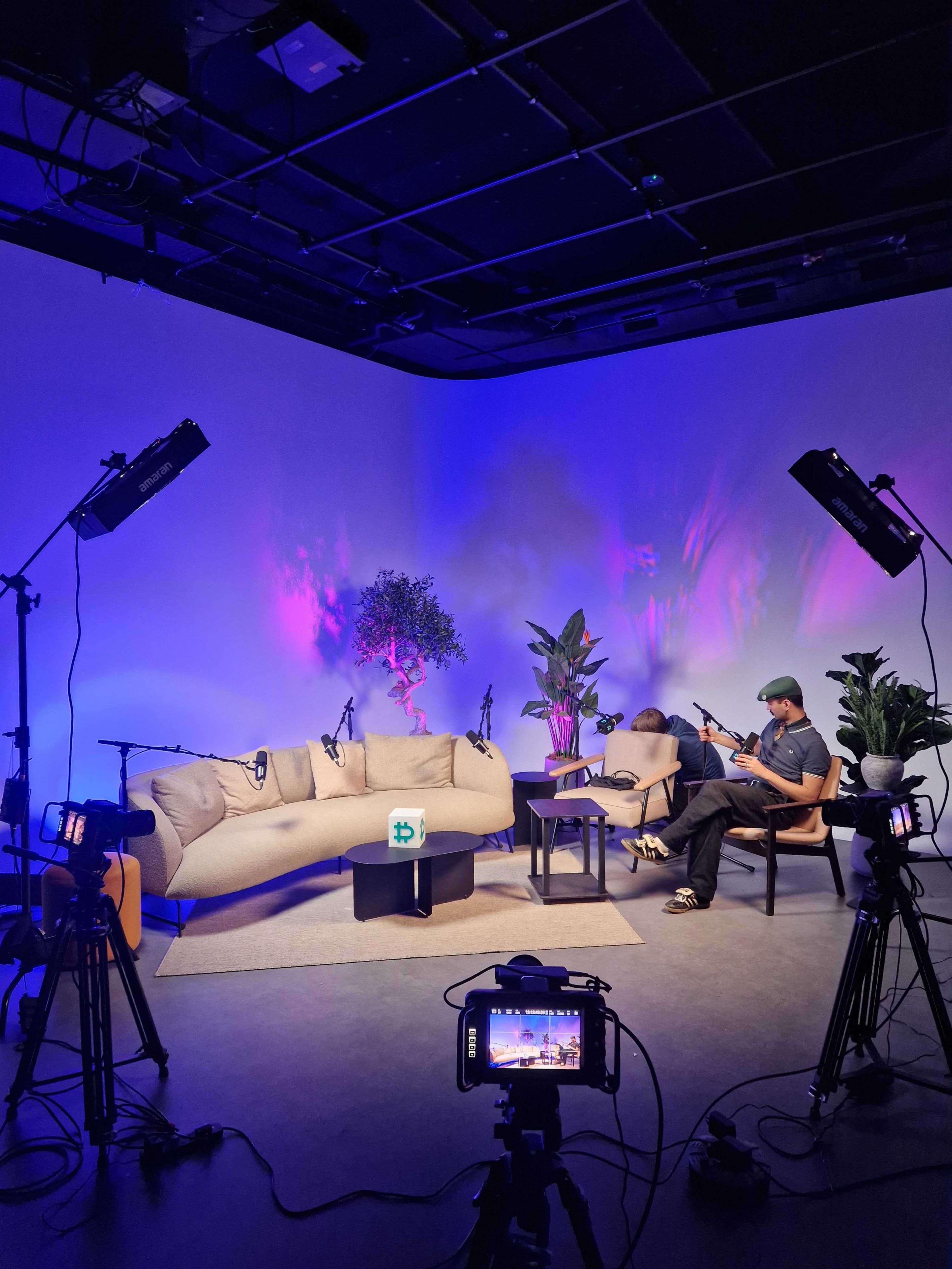 The image shows a studio setup with a beige couch, chairs, plants, and multiple cameras and lights arranged for a recorded discussion or interview.