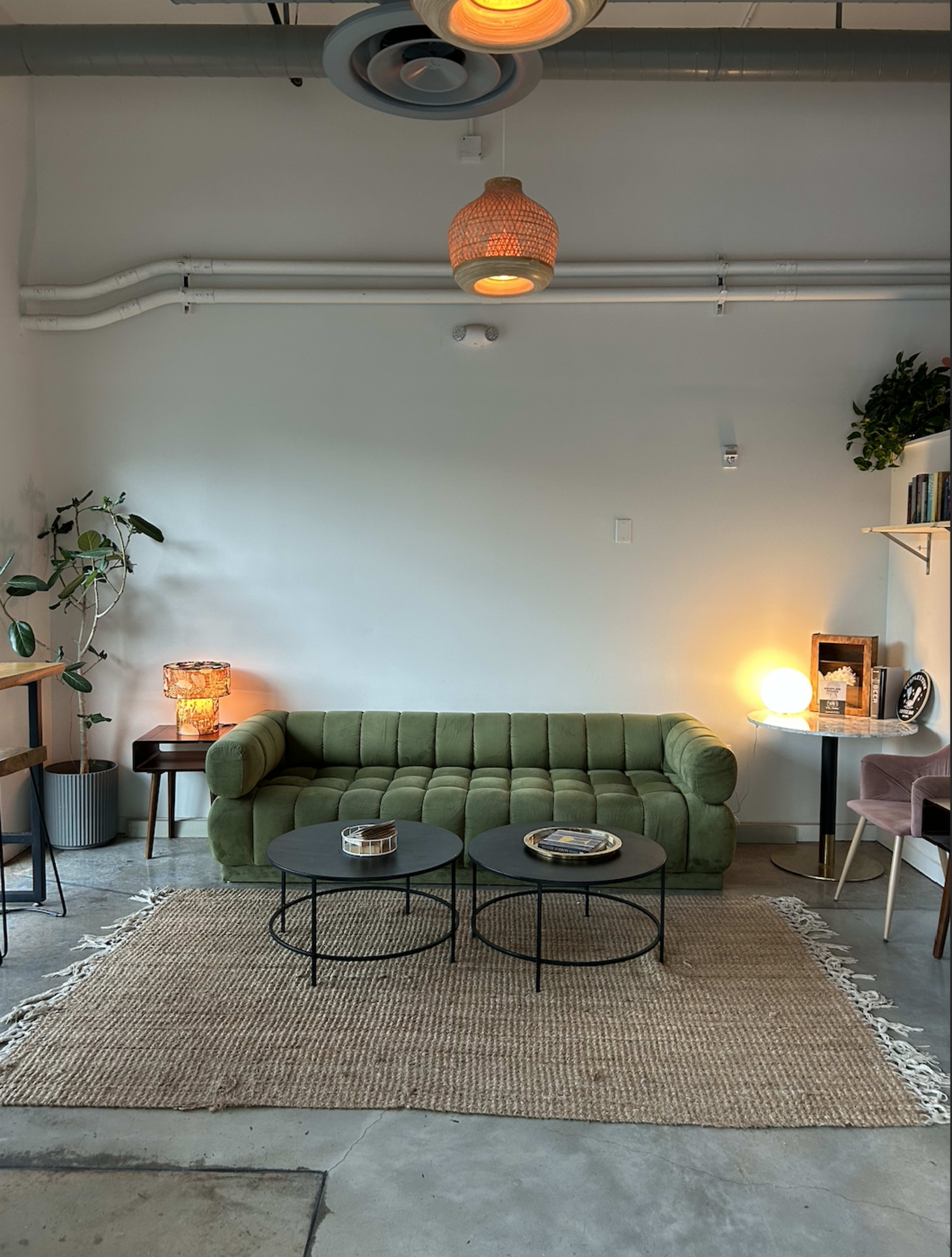 The image shows a cozy living room with a green tufted sofa, two round coffee tables, and various decorative elements including lamps and plants.