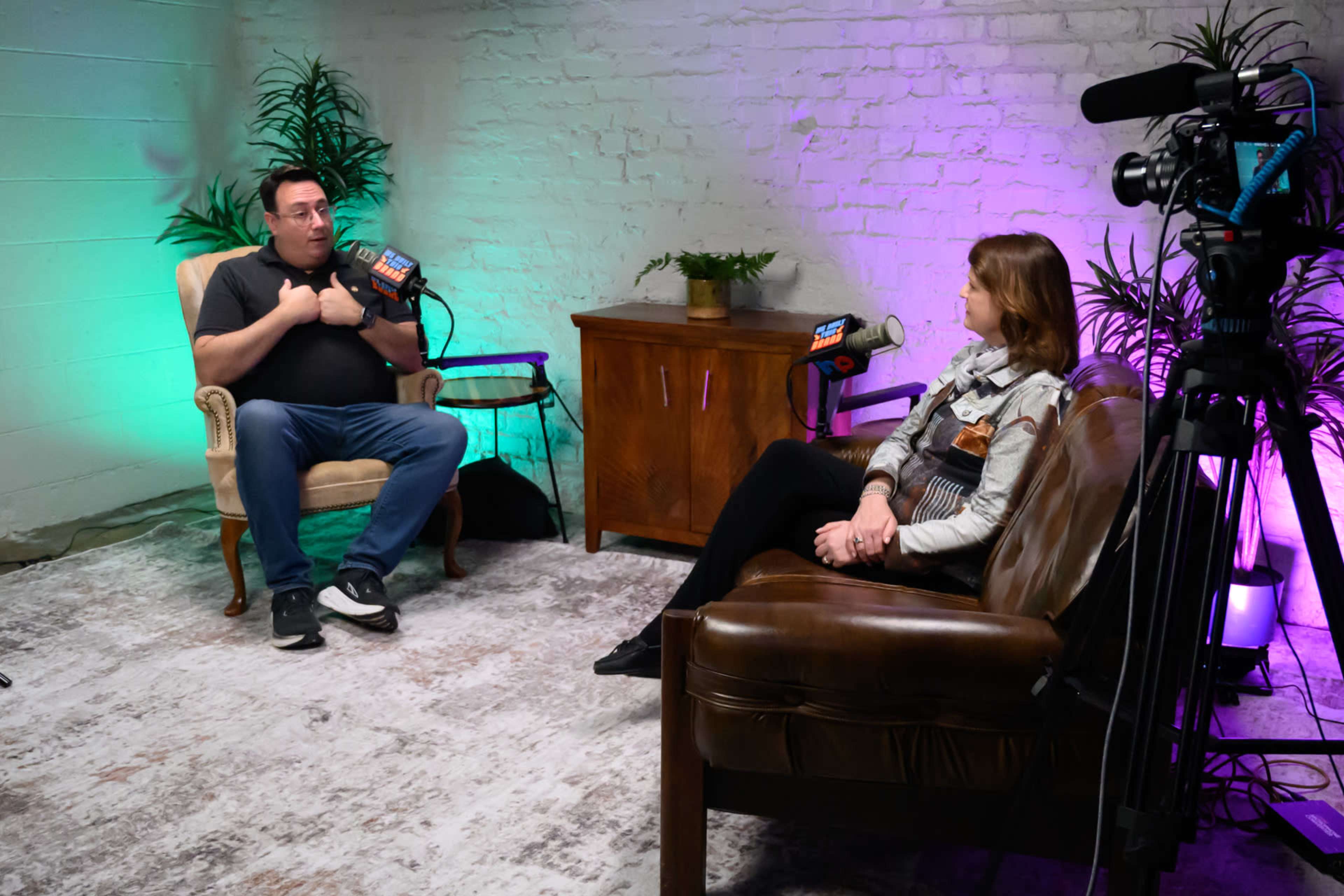 A man and a woman are engaged in a conversation in a softly lit studio, surrounded by potted plants and equipped with microphones and cameras.