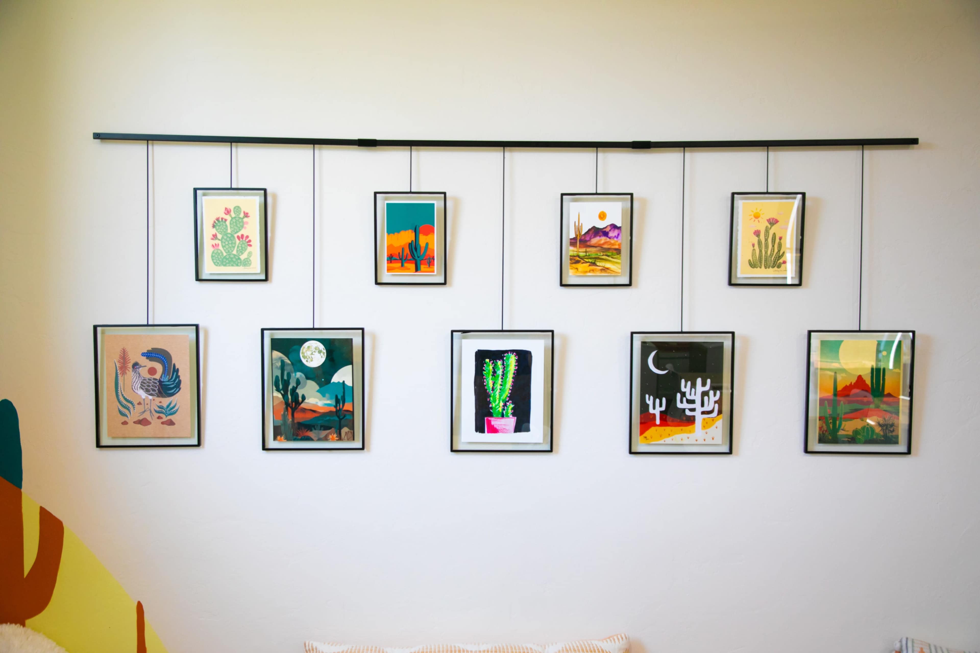 A series of framed artworks depicting desert landscapes and cacti are arranged on a wall with a horizontal hanging rod.