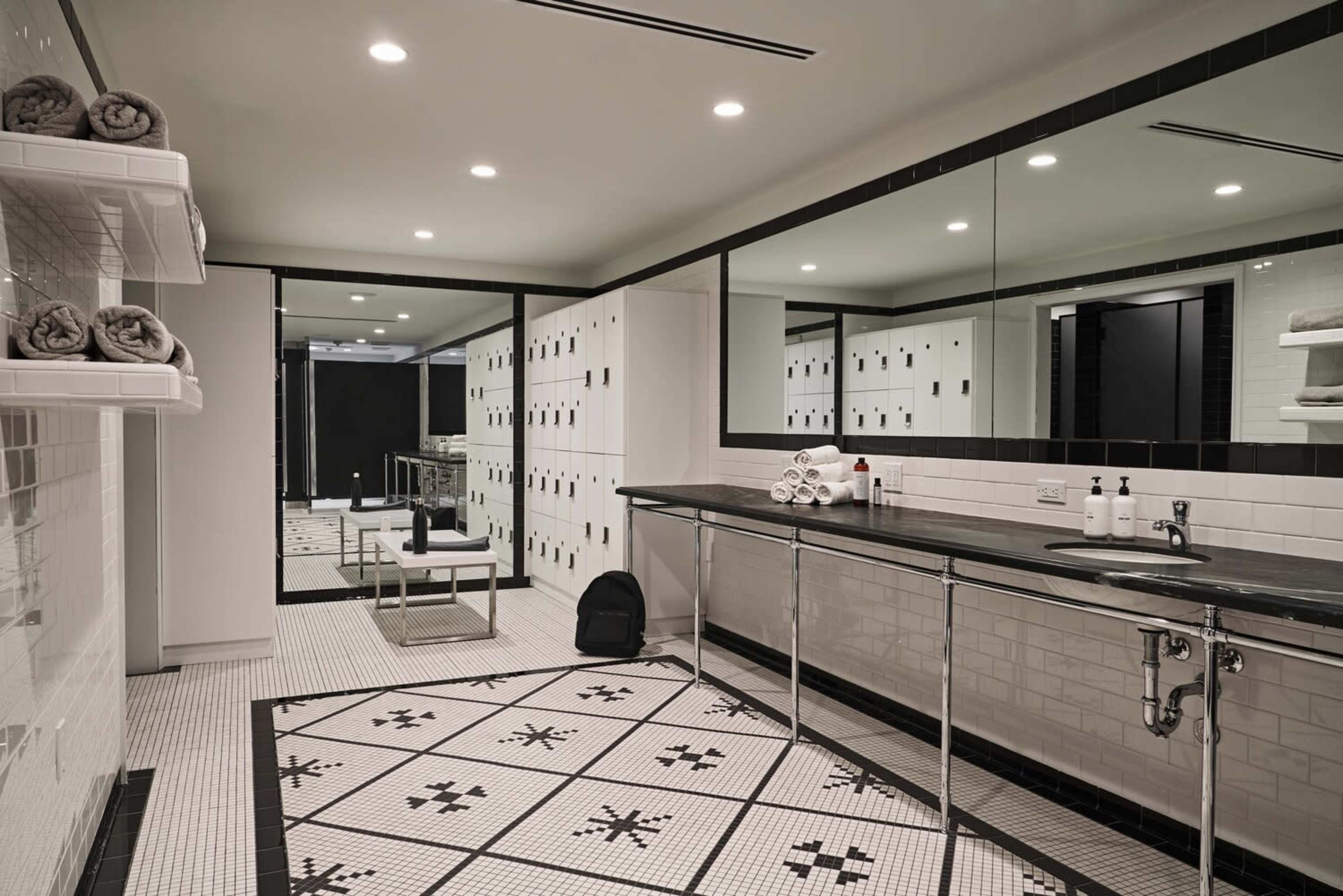 The image shows a modern bathroom with black and white tiled floors, a row of lockers, and a long countertop with toiletries and neatly rolled towels.