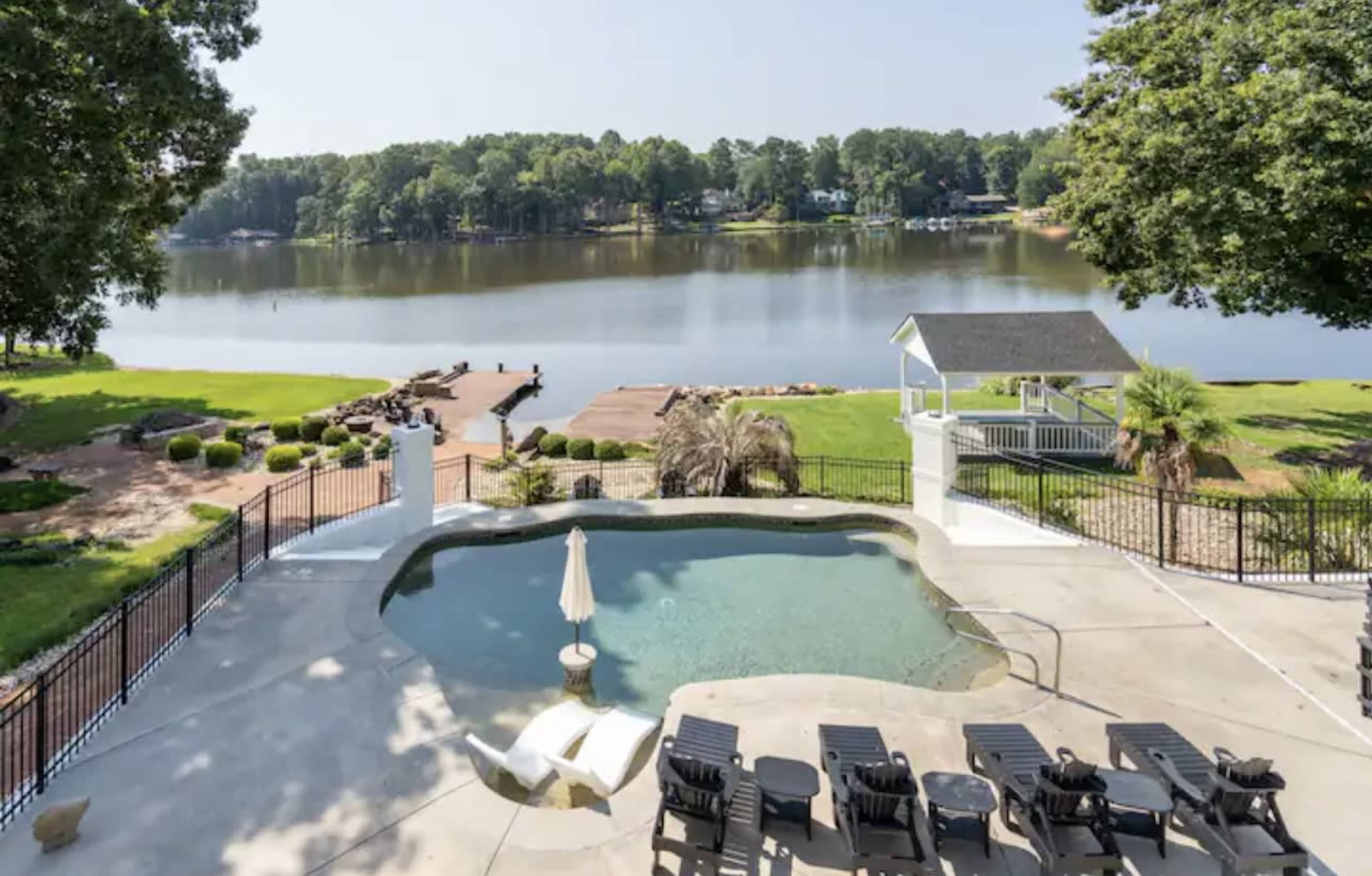 The image shows a backyard pool with lounge chairs, overlooking a calm lake surrounded by trees and a small dock.