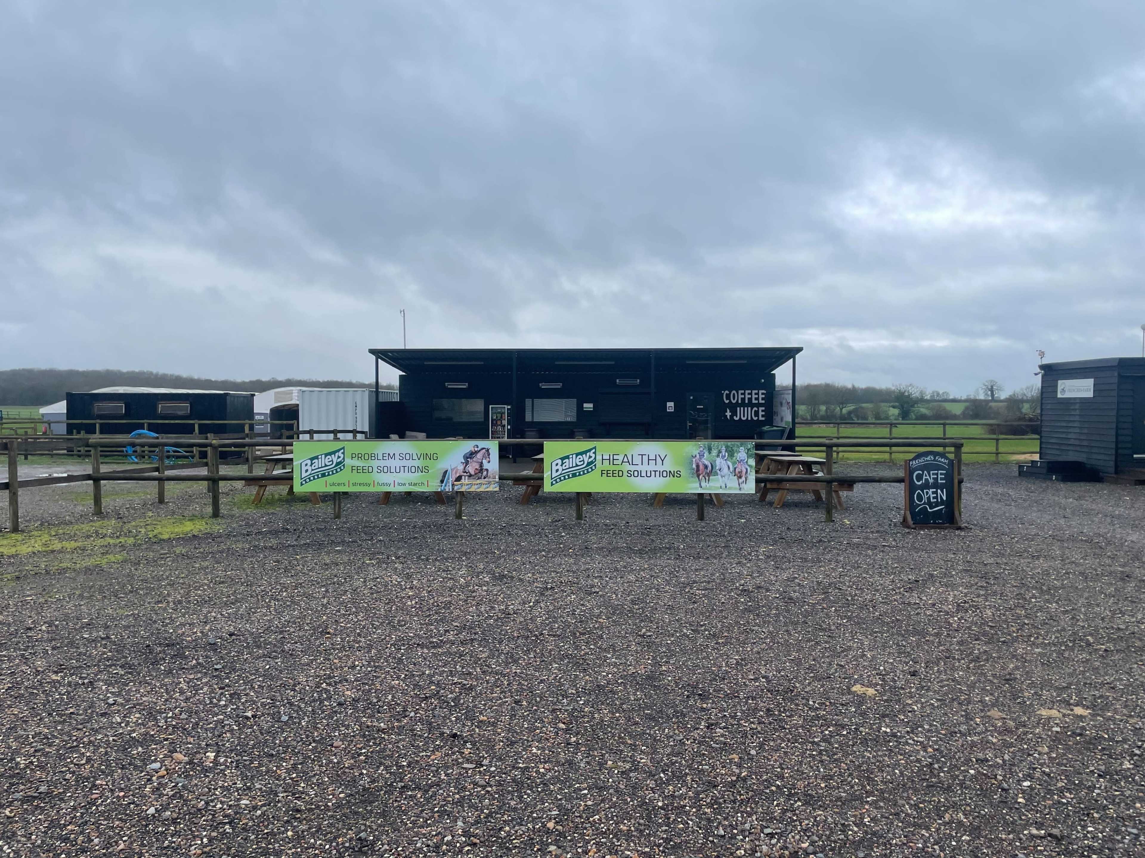A small outdoor café with a service window and picnic tables is situated in a gravel area, surrounded by open fields under a cloudy sky.