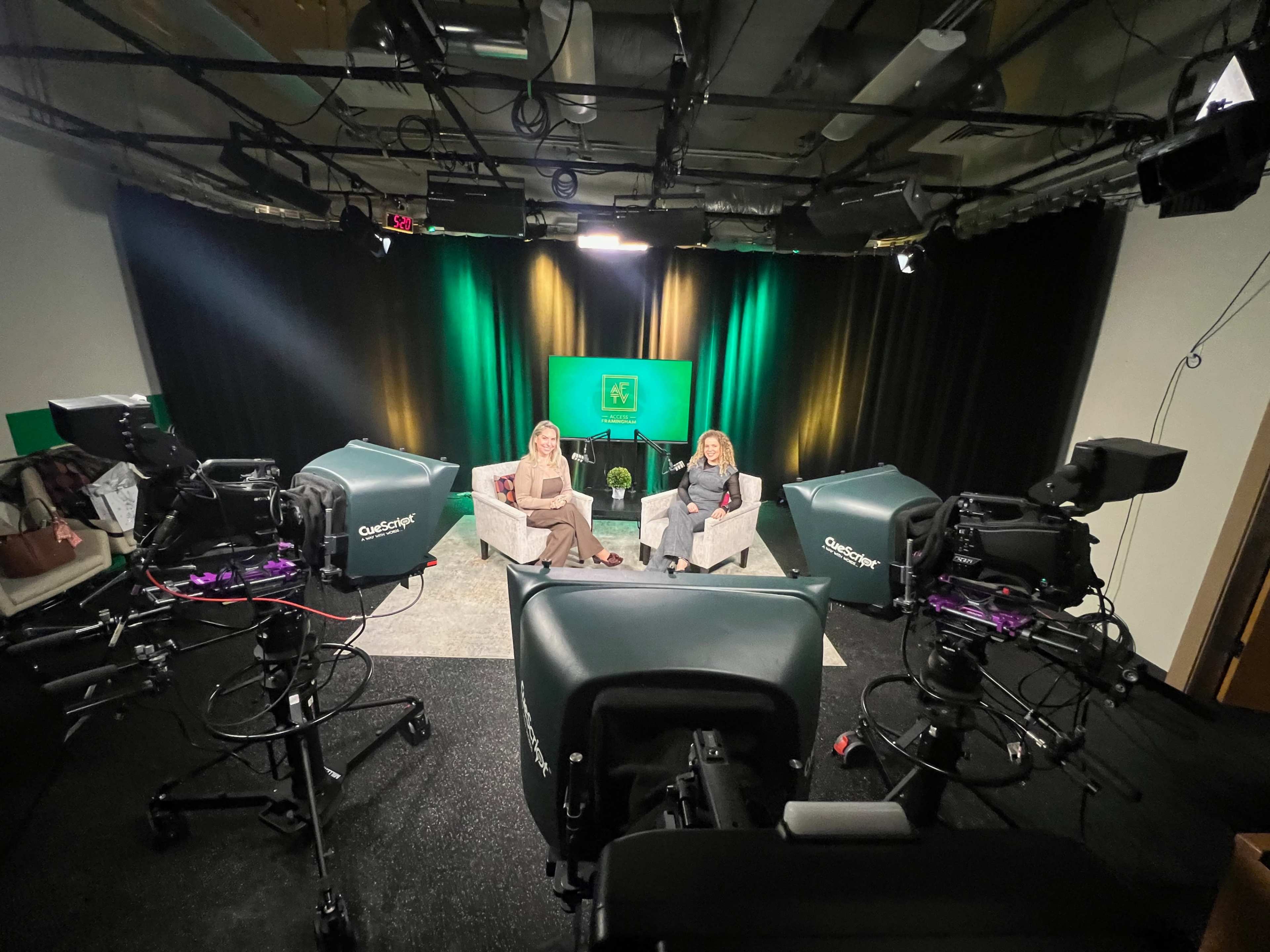The image shows a television studio setup with two women seated on couches, flanked by camera equipment and a backdrop featuring a green logo.