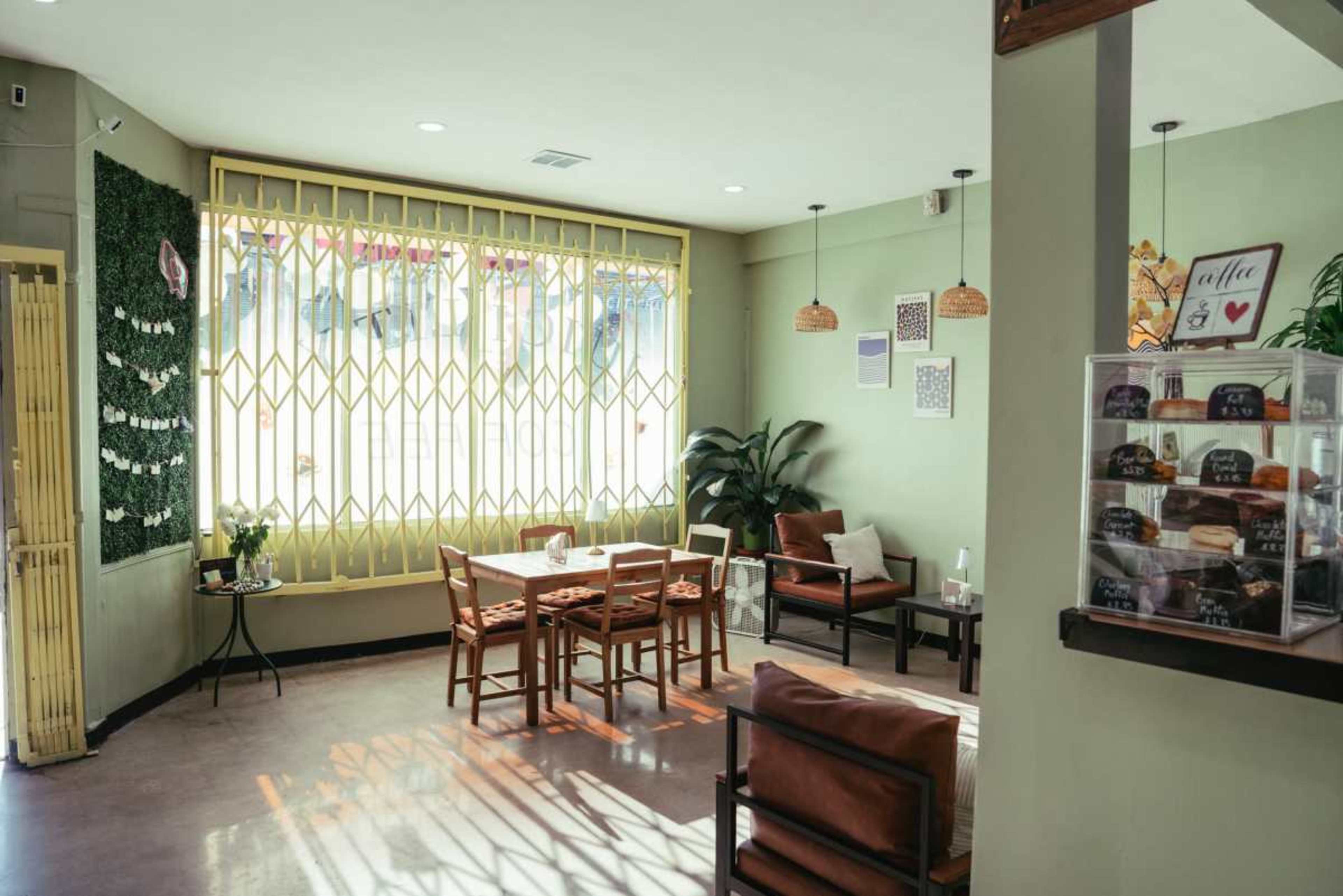 The image shows a cozy cafe interior with wooden furniture, green walls, and large windows letting in natural light.