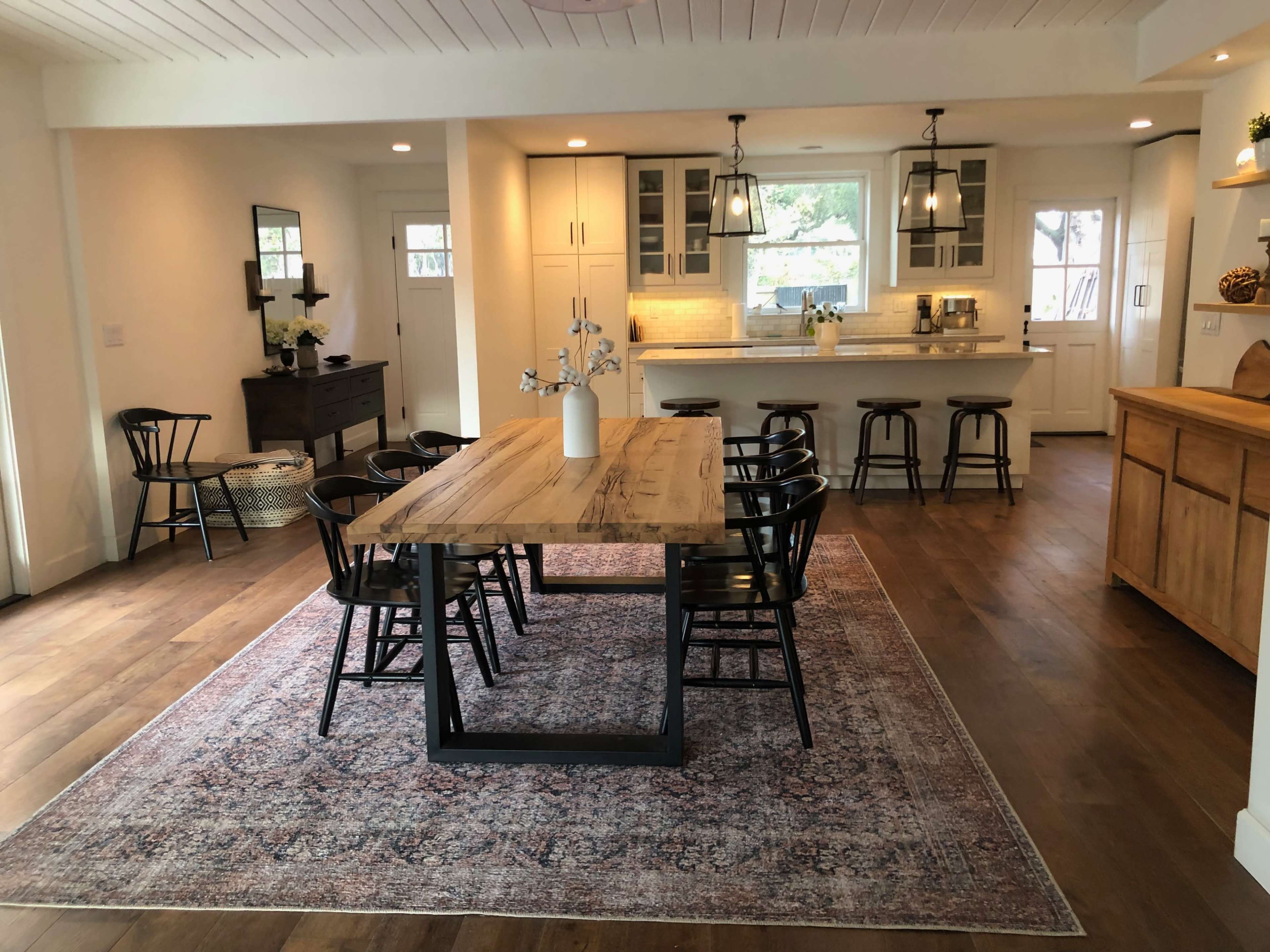 A dining area with a large wooden table surrounded by black chairs, set on a patterned rug, adjacent to a kitchen featuring light cabinetry and pendant lighting.
