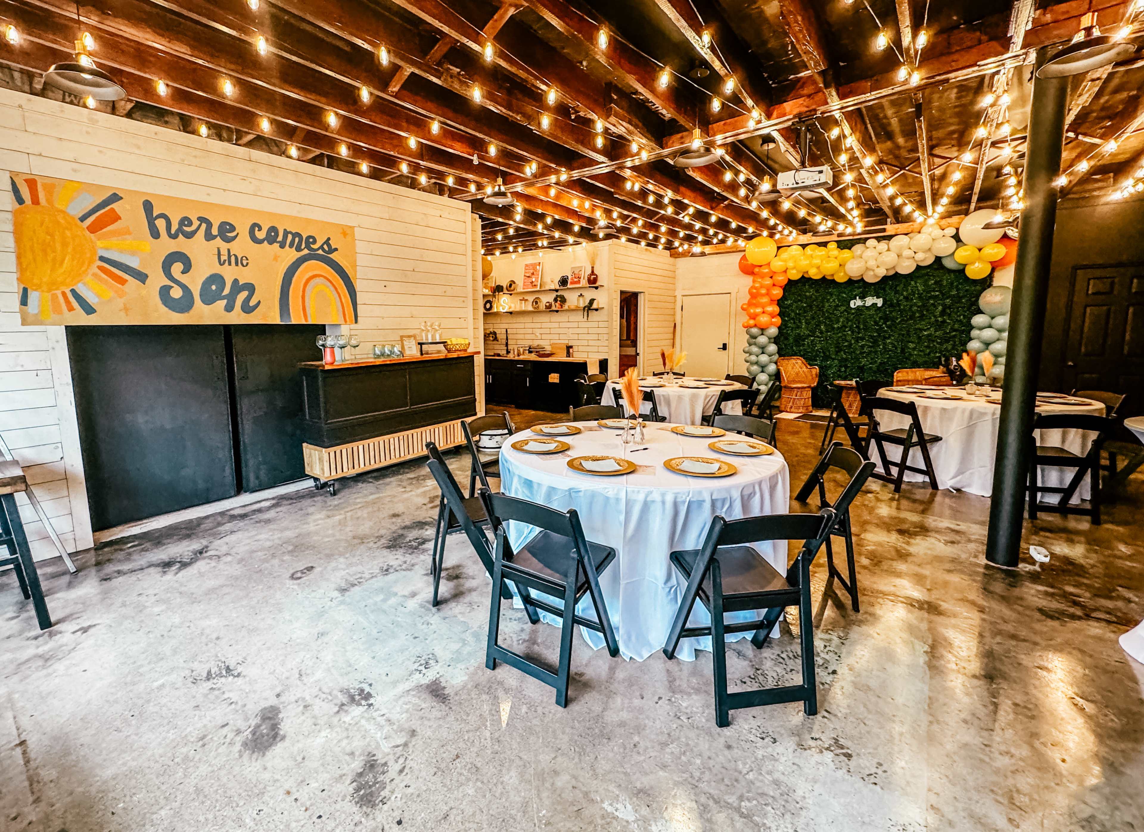 A decorated event space with round tables set up, a colorful mural on the wall, and strings of lights hanging from the ceiling.