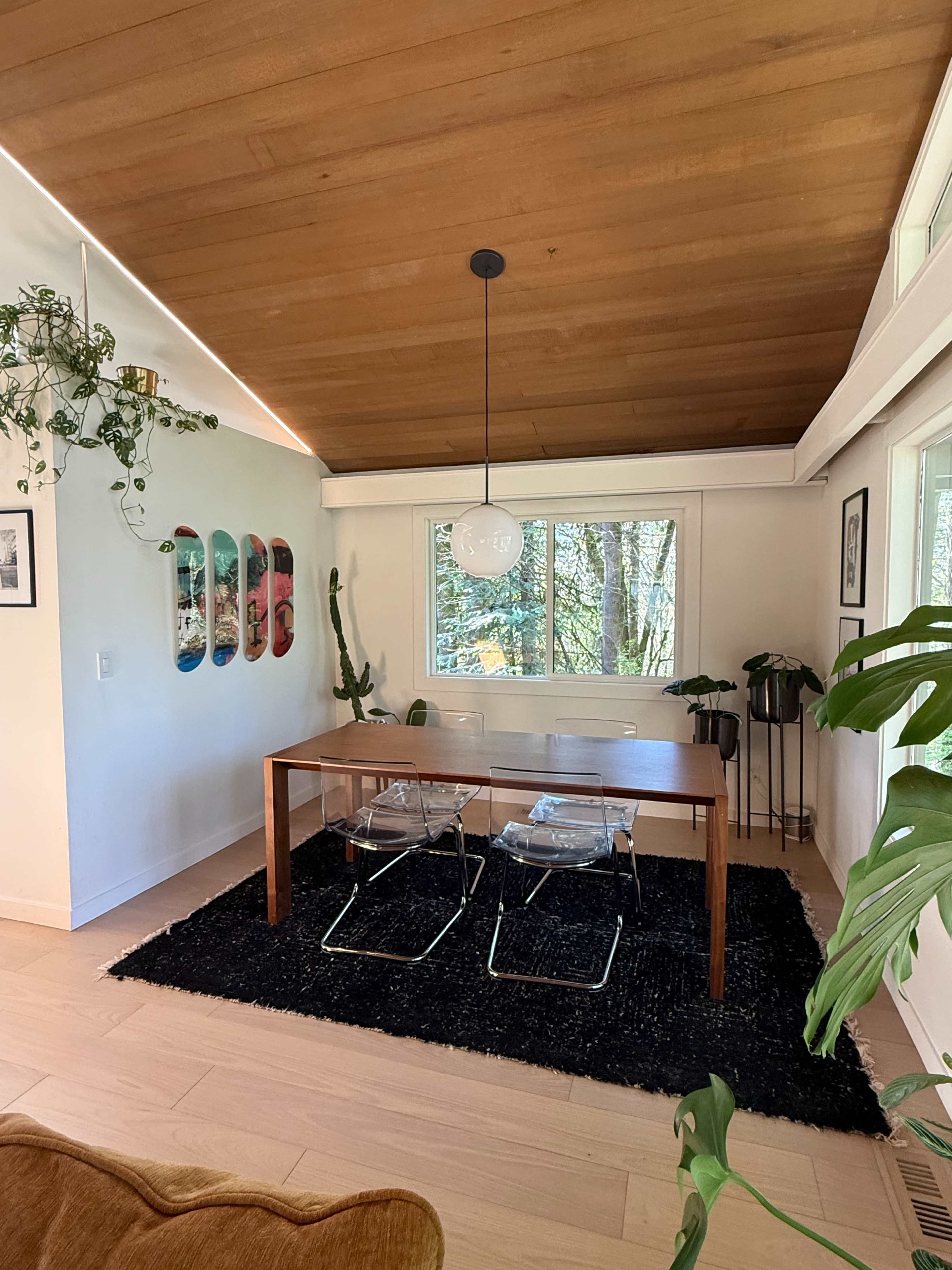 The image shows a bright dining area with a wooden table, two clear chairs, and a black rug, surrounded by plants and artwork on the walls.