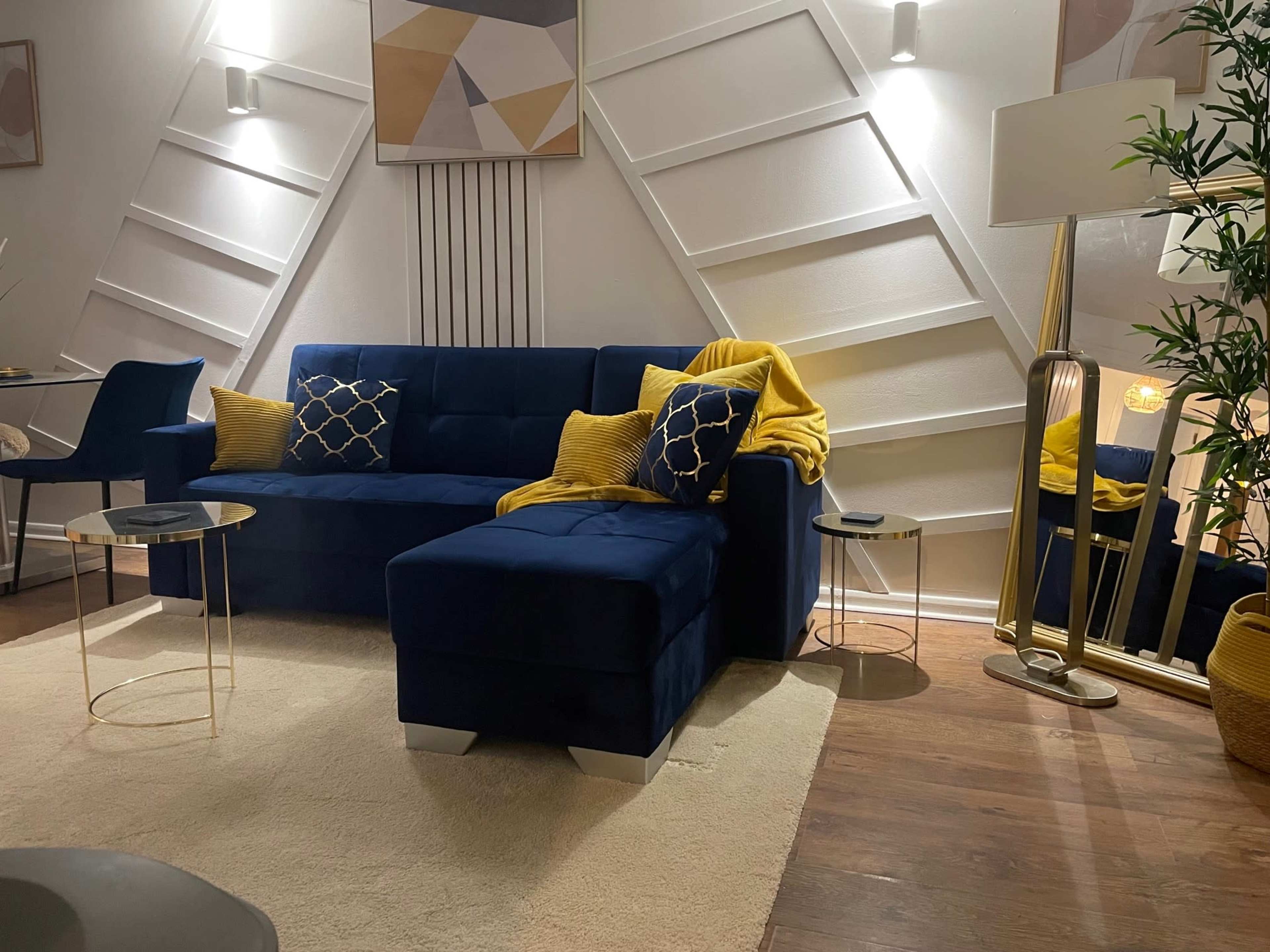 The image shows a modern living room featuring a navy blue sectional sofa with decorative pillows, a yellow throw blanket, two small round side tables, and a standing lamp next to a potted plant.