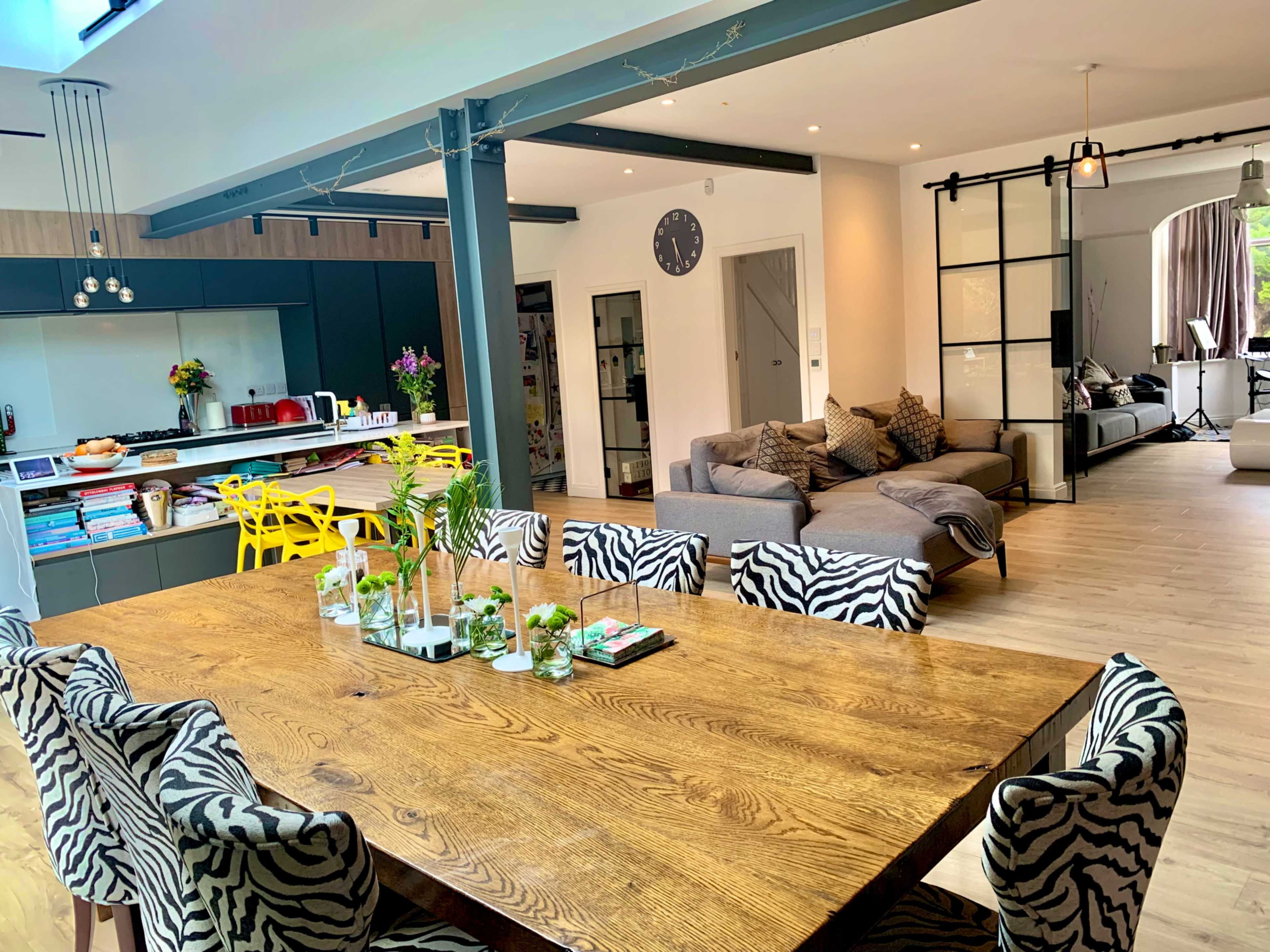 A modern open-plan living space featuring a large wooden dining table with zebra-print chairs, a sleek kitchen area with a breakfast bar, and a cozy seating area with a sectional couch.