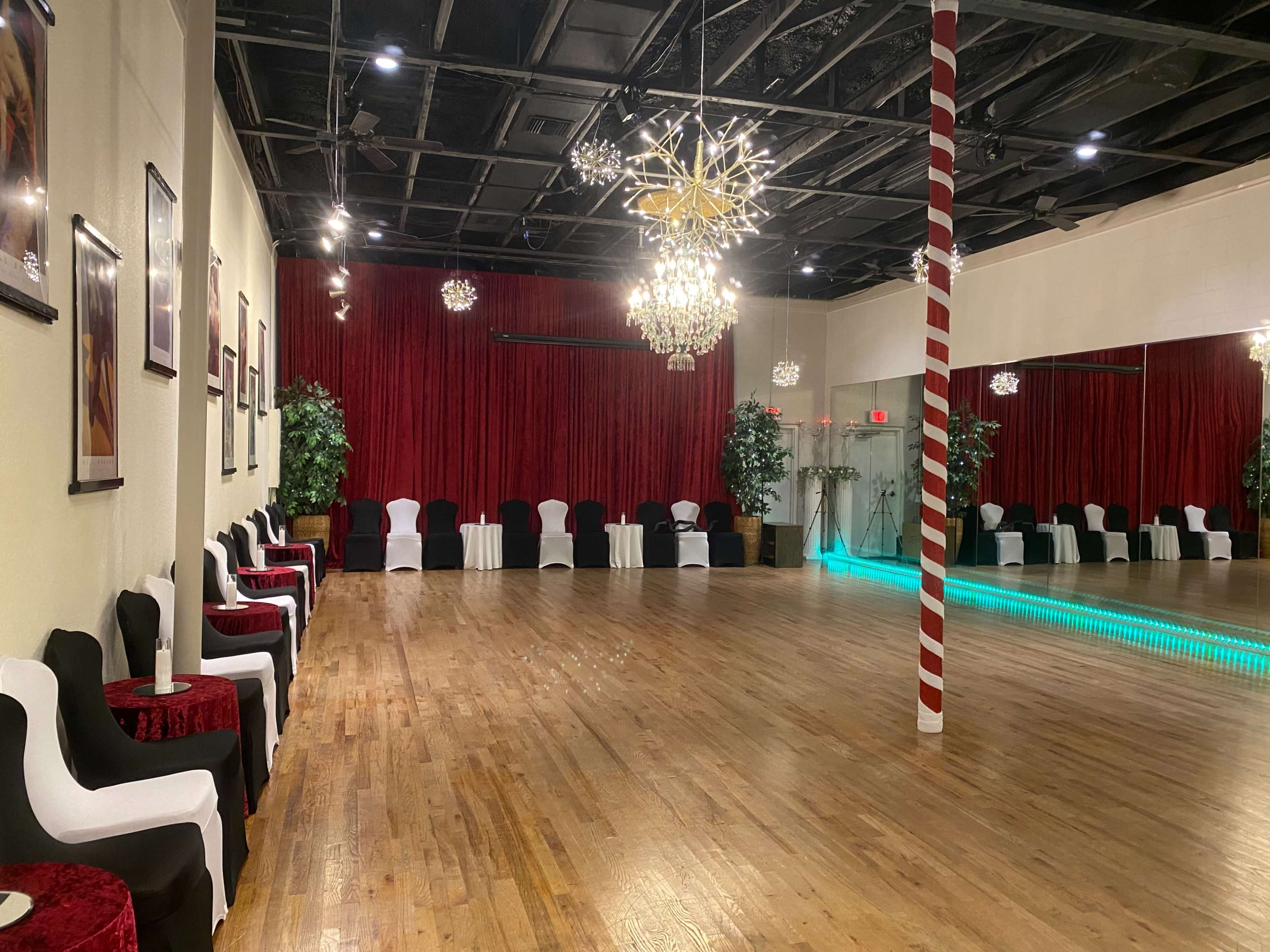 A spacious dance studio with a wooden floor, red curtains, and decorative lighting, including chandeliers and a row of chairs along the walls.