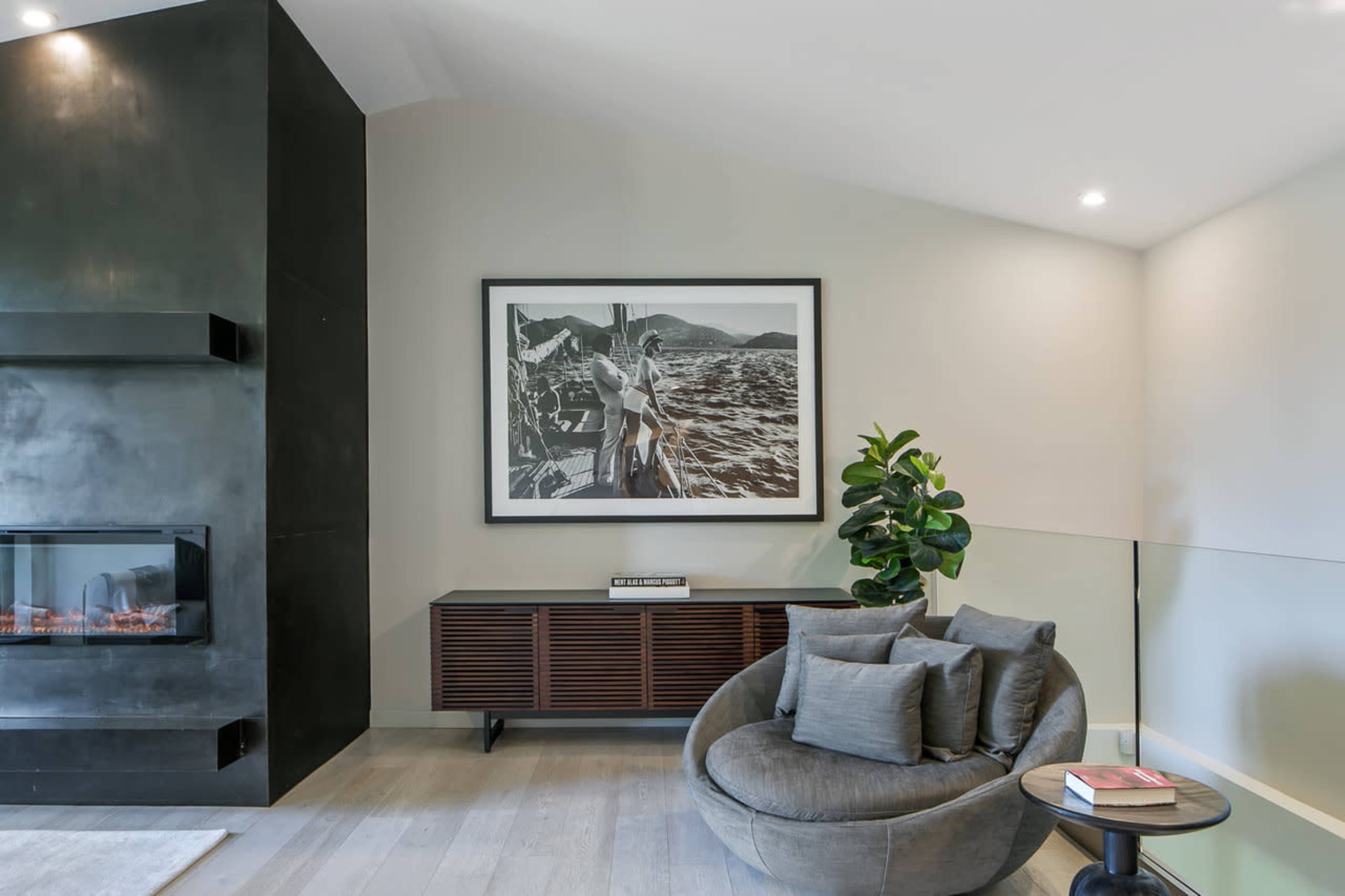 A modern living room features a circular chair, a low wooden sideboard, a large black-and-white framed photograph, and a wall-mounted fireplace.