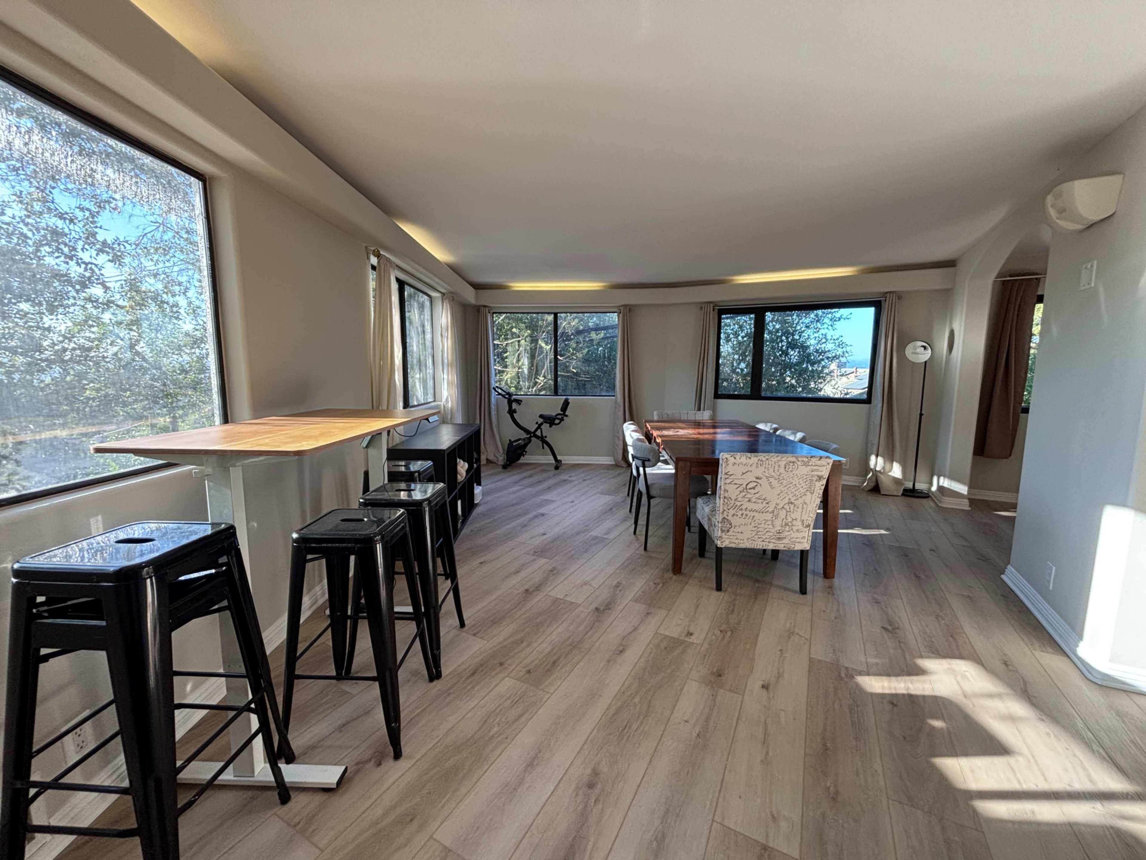 The image shows a modern room with large windows, a dining table, black bar stools, and a stationary bike.