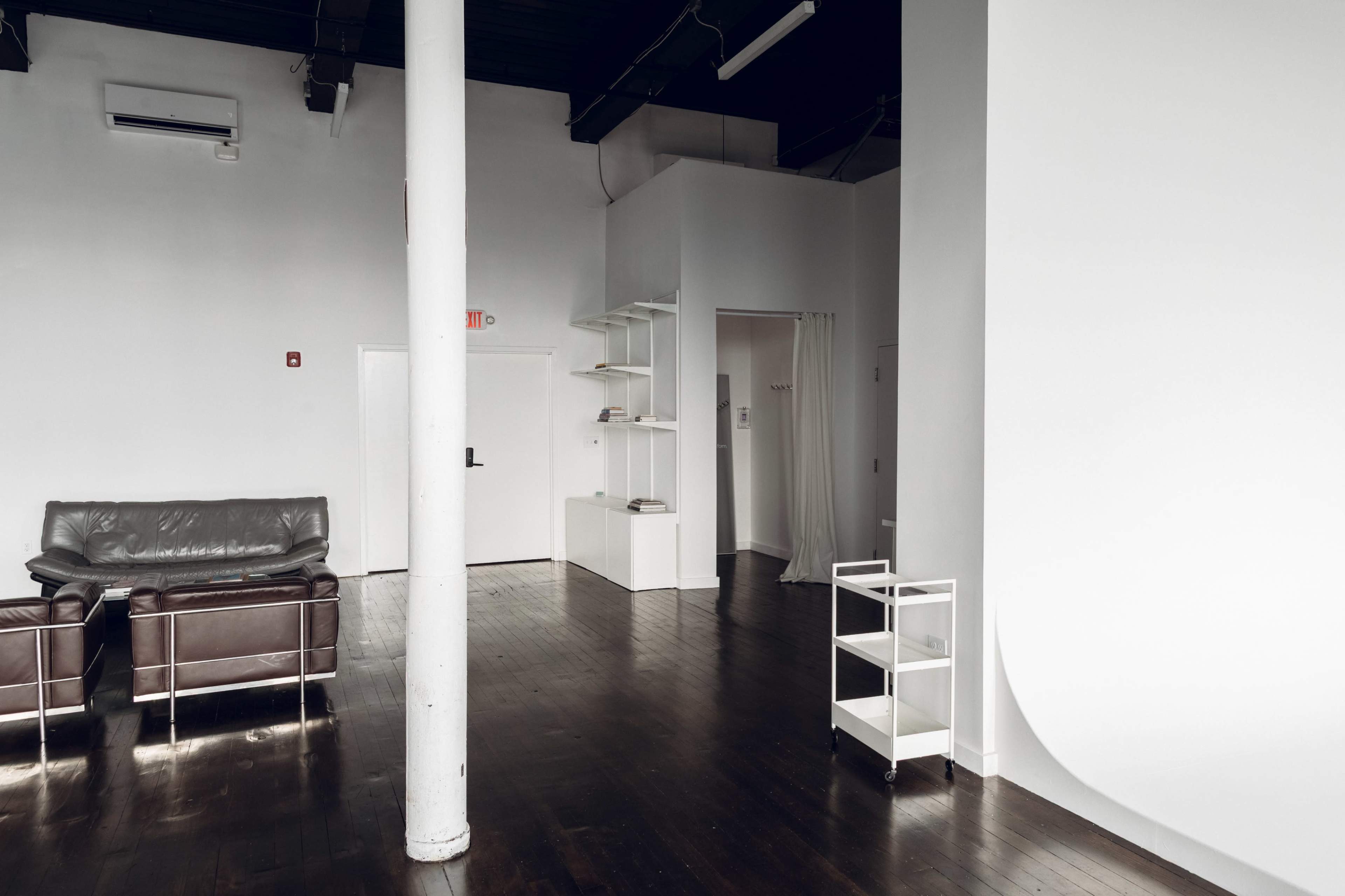 The image shows a spacious, minimalistic interior with dark wooden floors, a leather couch, and a white wall featuring a small bookshelf and a doorway.