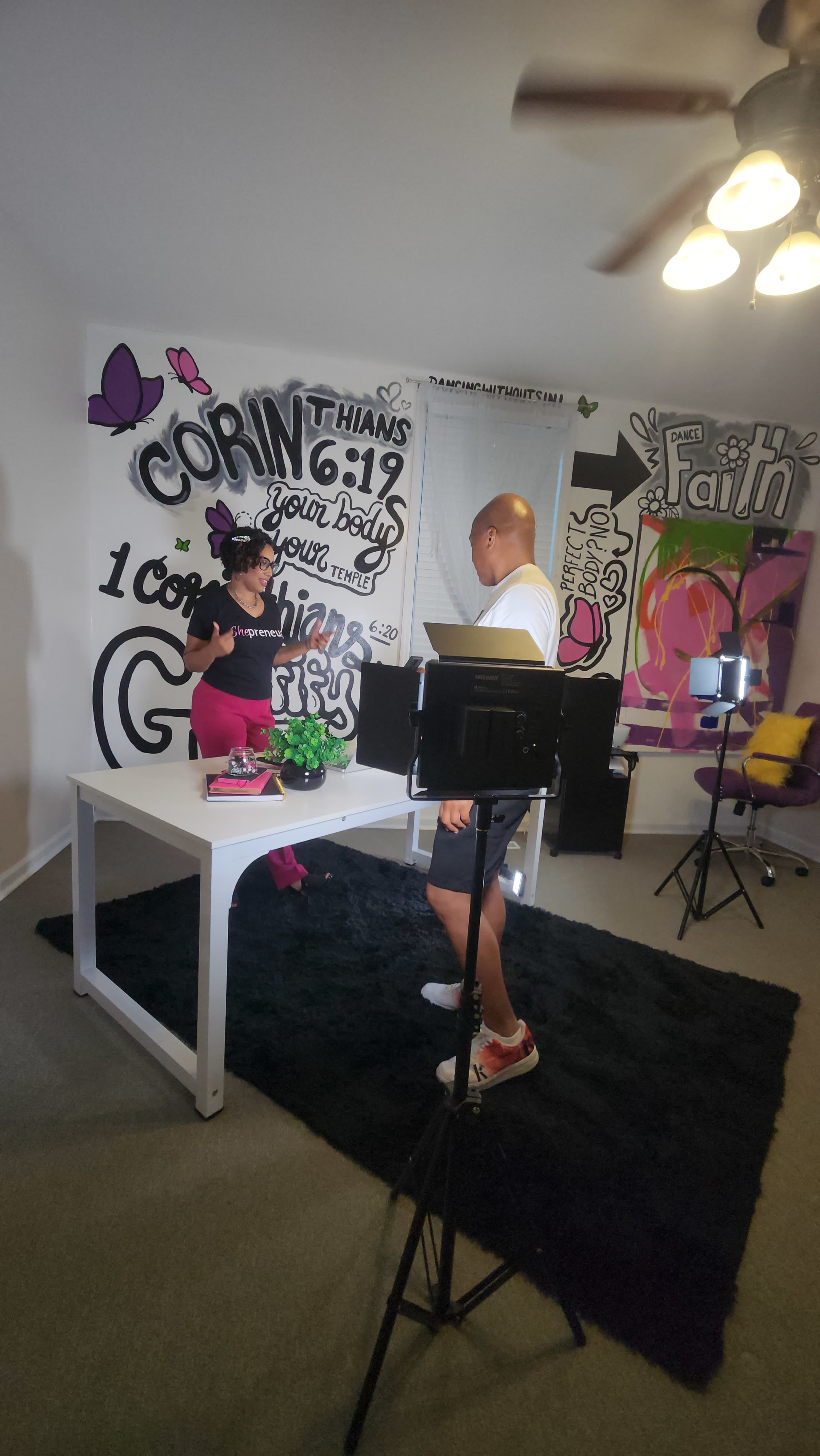 A woman stands at a table with plants while a man, facing her, is positioned in a room decorated with vibrant wall art and equipped with lighting and cameras.