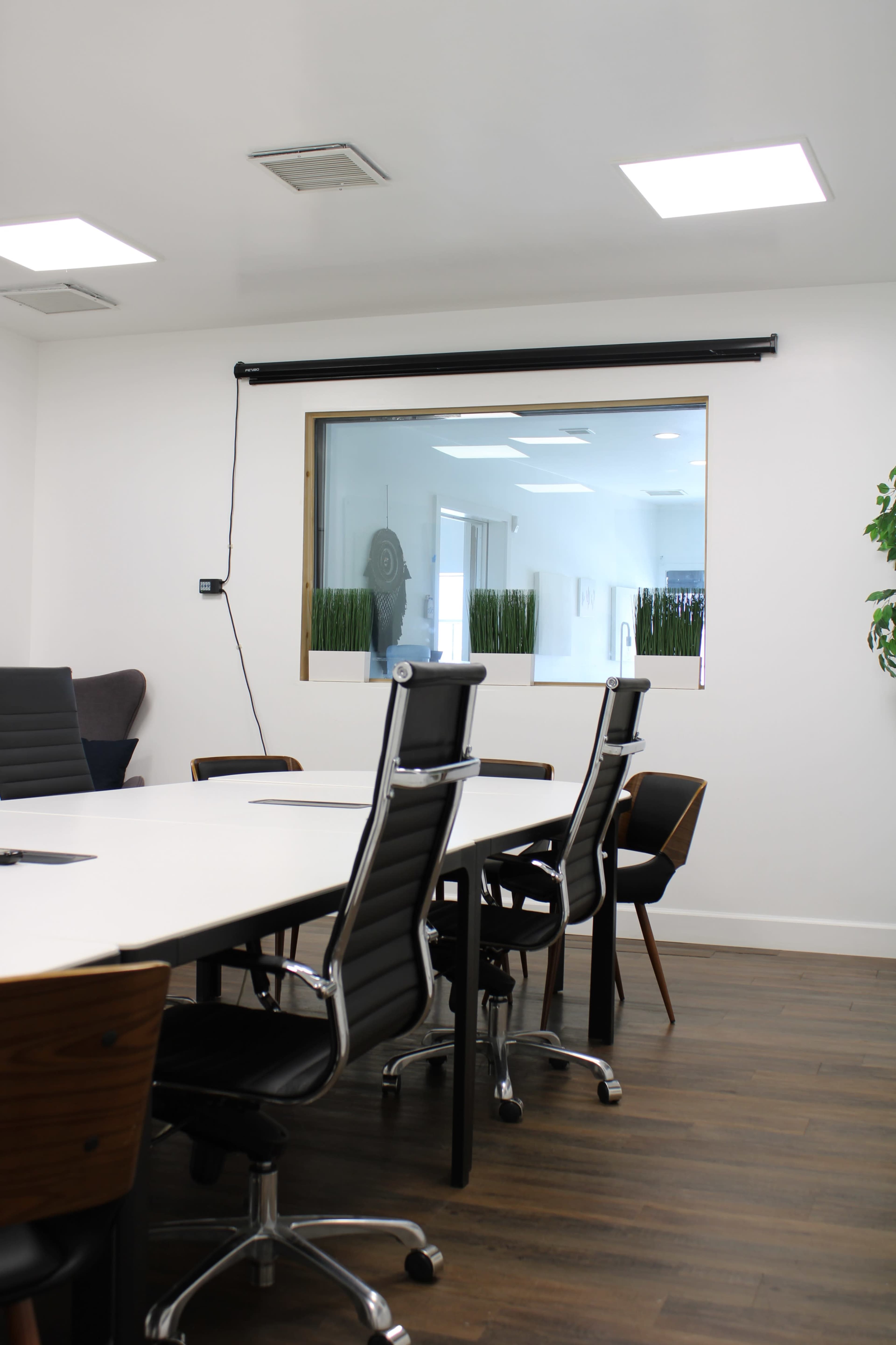 A modern conference room features a large table surrounded by black and wooden chairs, with a window overlooking a bright, plant-filled area.