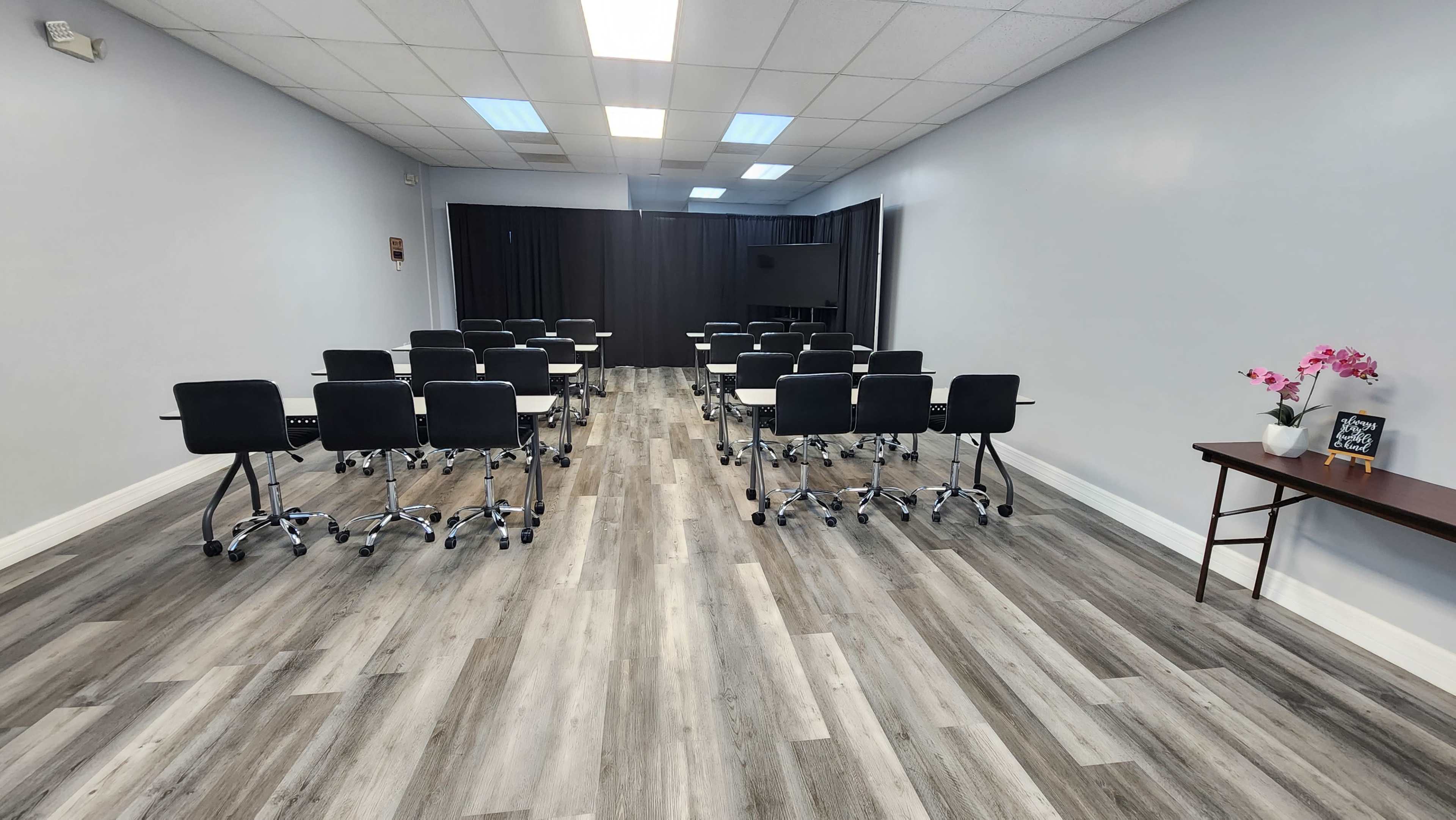 A modern meeting room features rows of black chairs on wheels facing a front area with a table and decorative flowers.