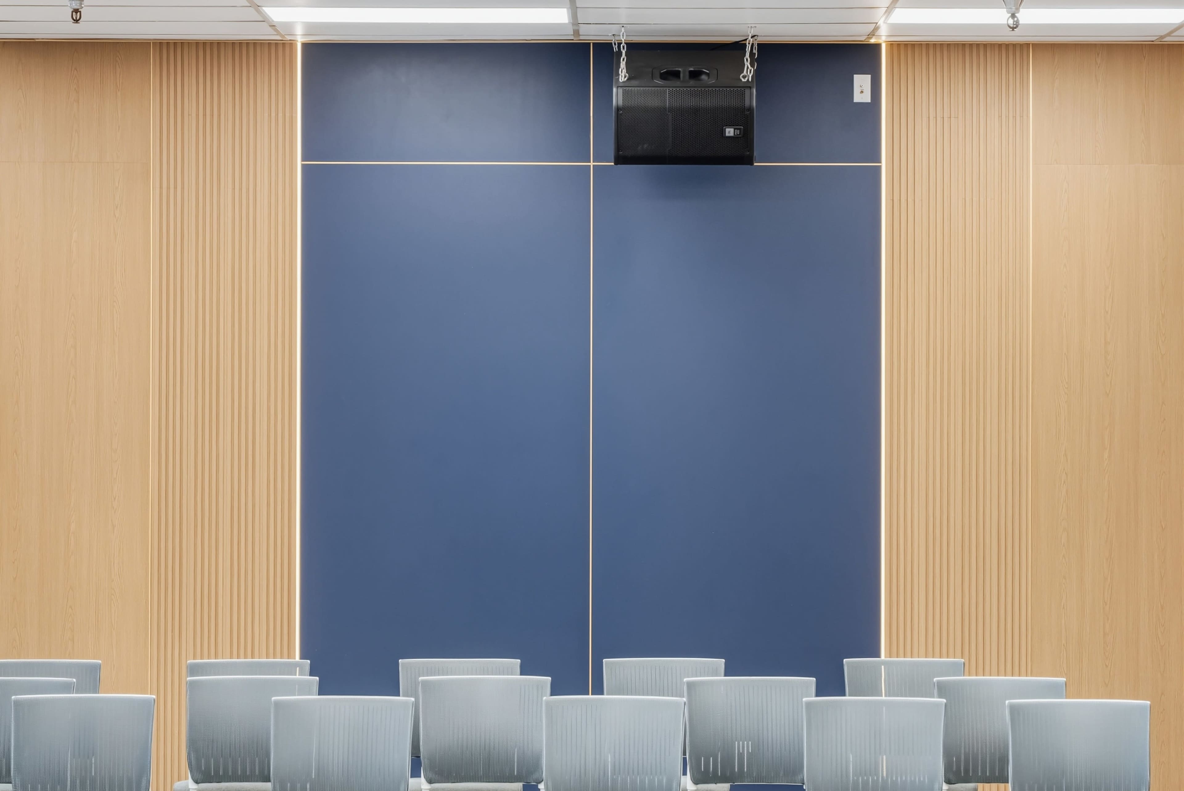 The image shows a classroom or conference room setup with rows of light-colored chairs facing a blue wall that features a mounted speaker.