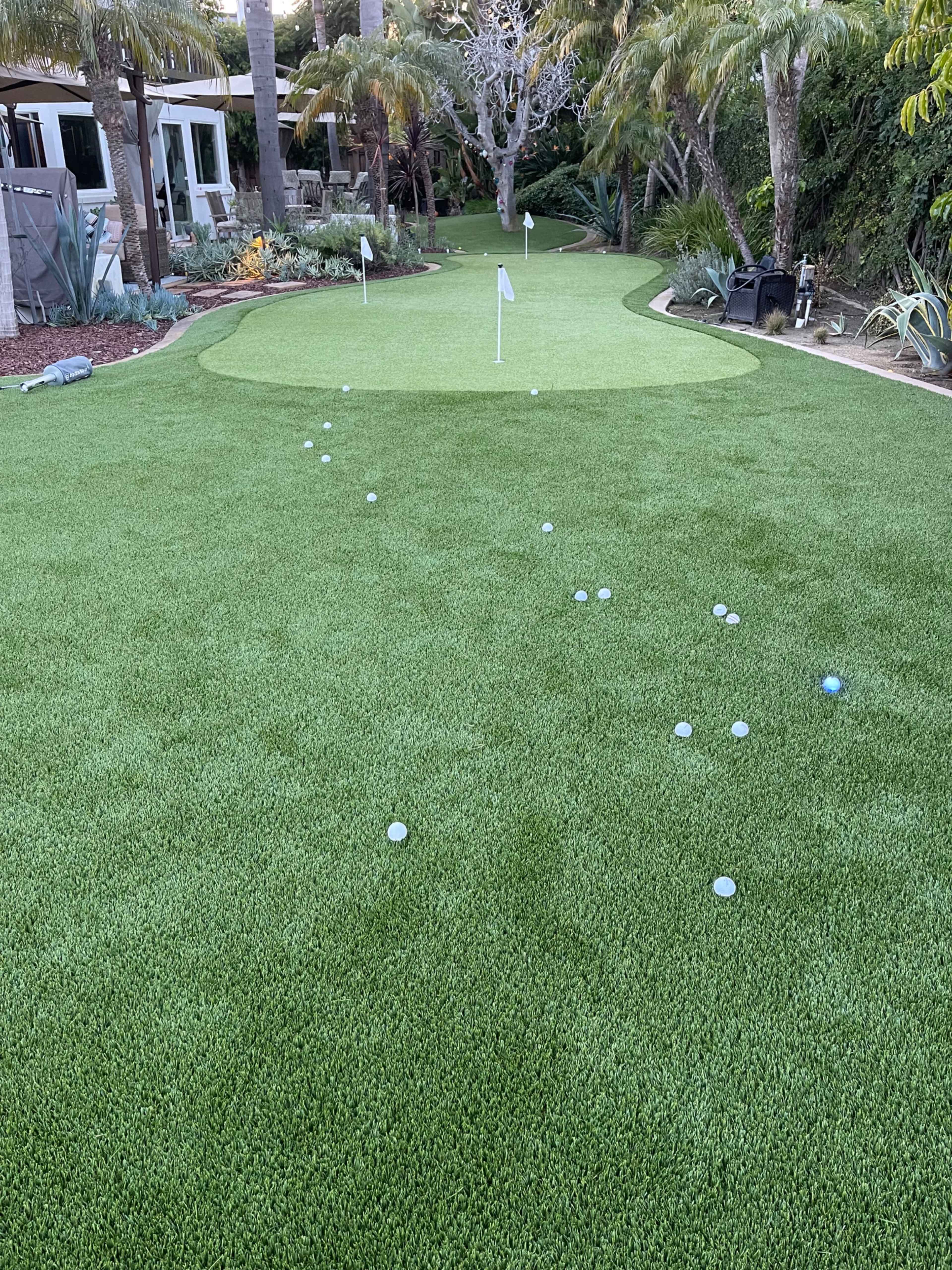 The image shows a well-maintained artificial putting green in a backyard, with multiple golf balls scattered around and a flag positioned on the green.
