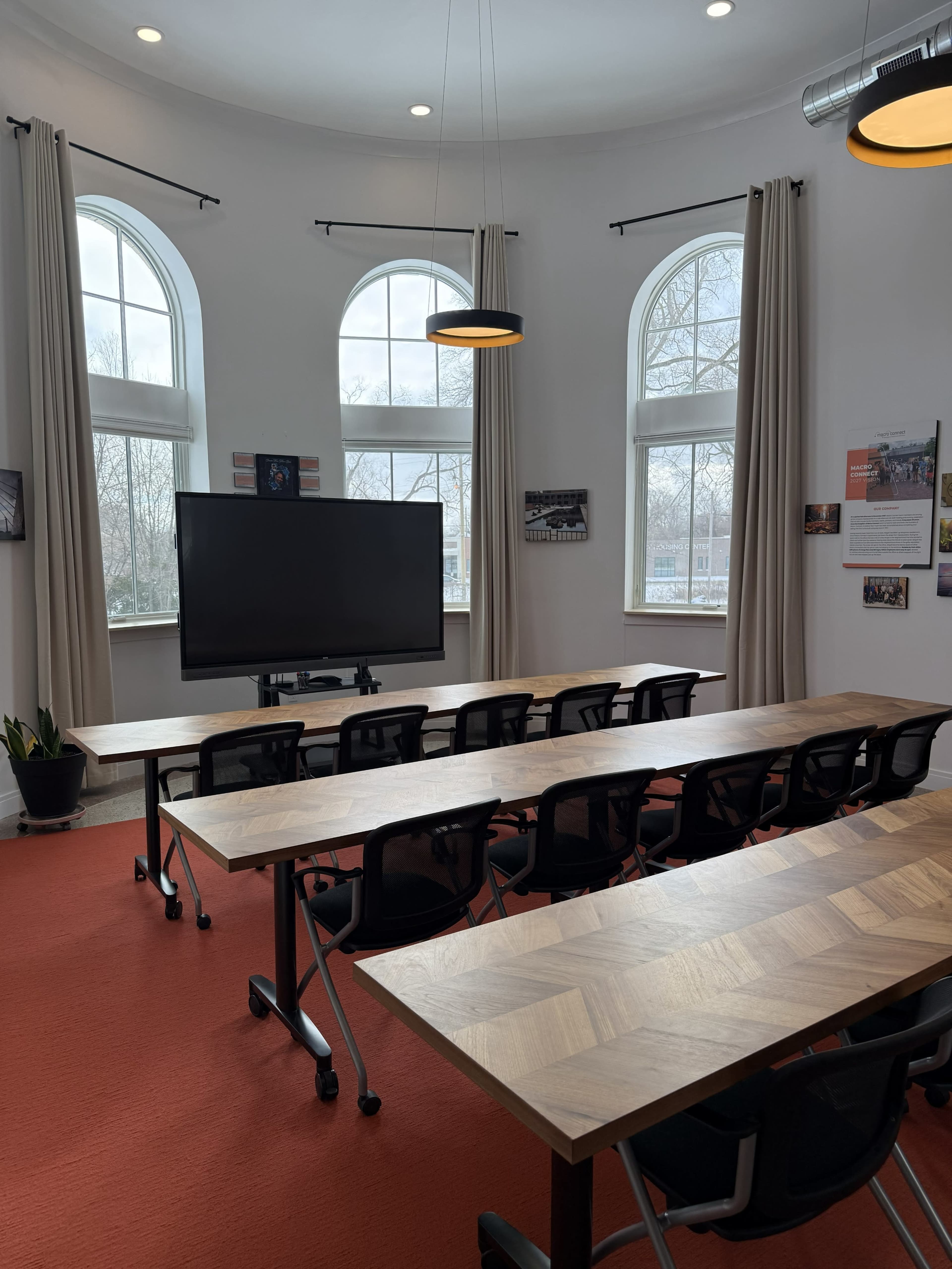 The image shows a spacious meeting room equipped with rows of tables and chairs, a large screen at the front, and large windows allowing natural light.