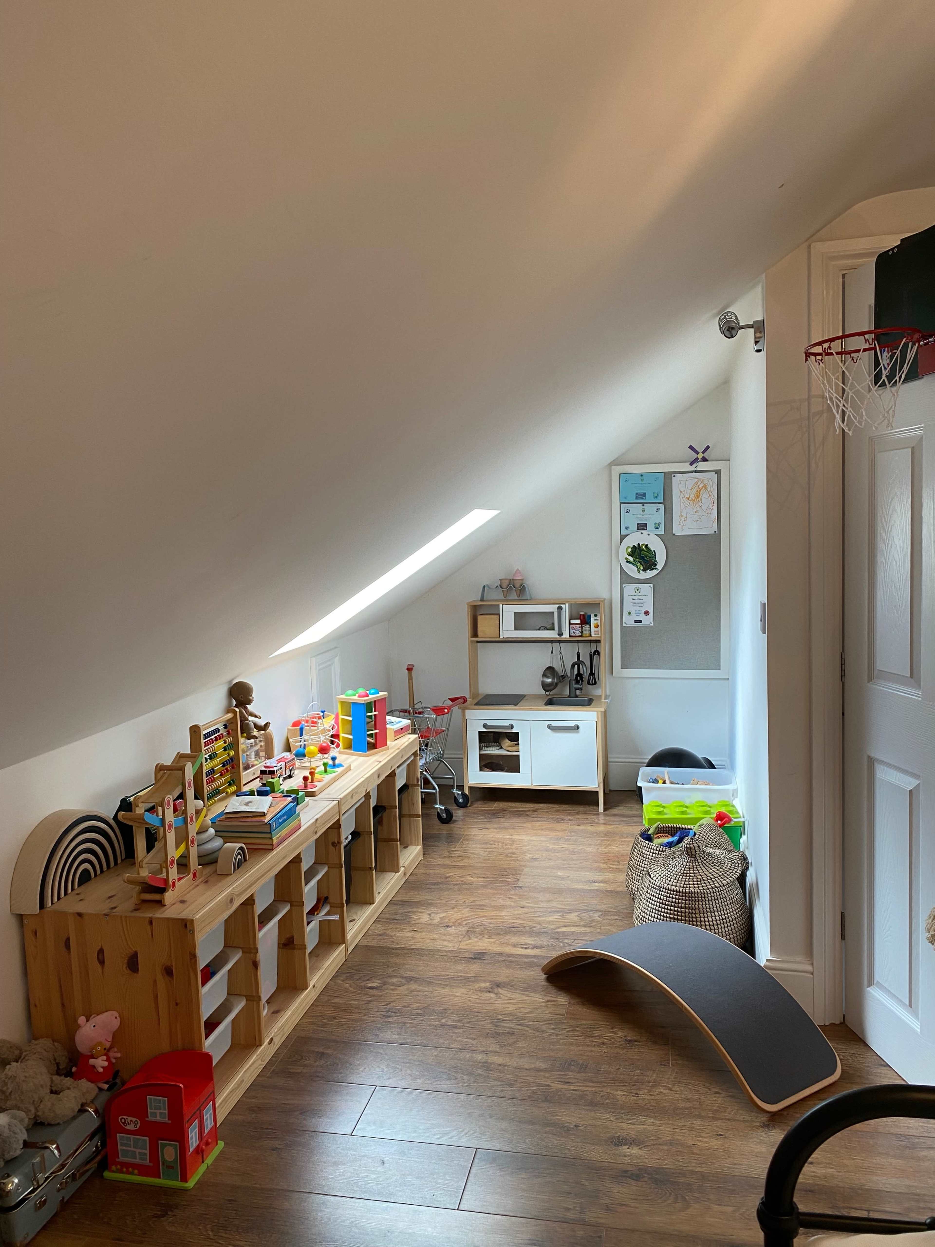 A slanted attic room features a play area with a shelving unit filled with toys, a small kitchen set, and a basketball hoop.