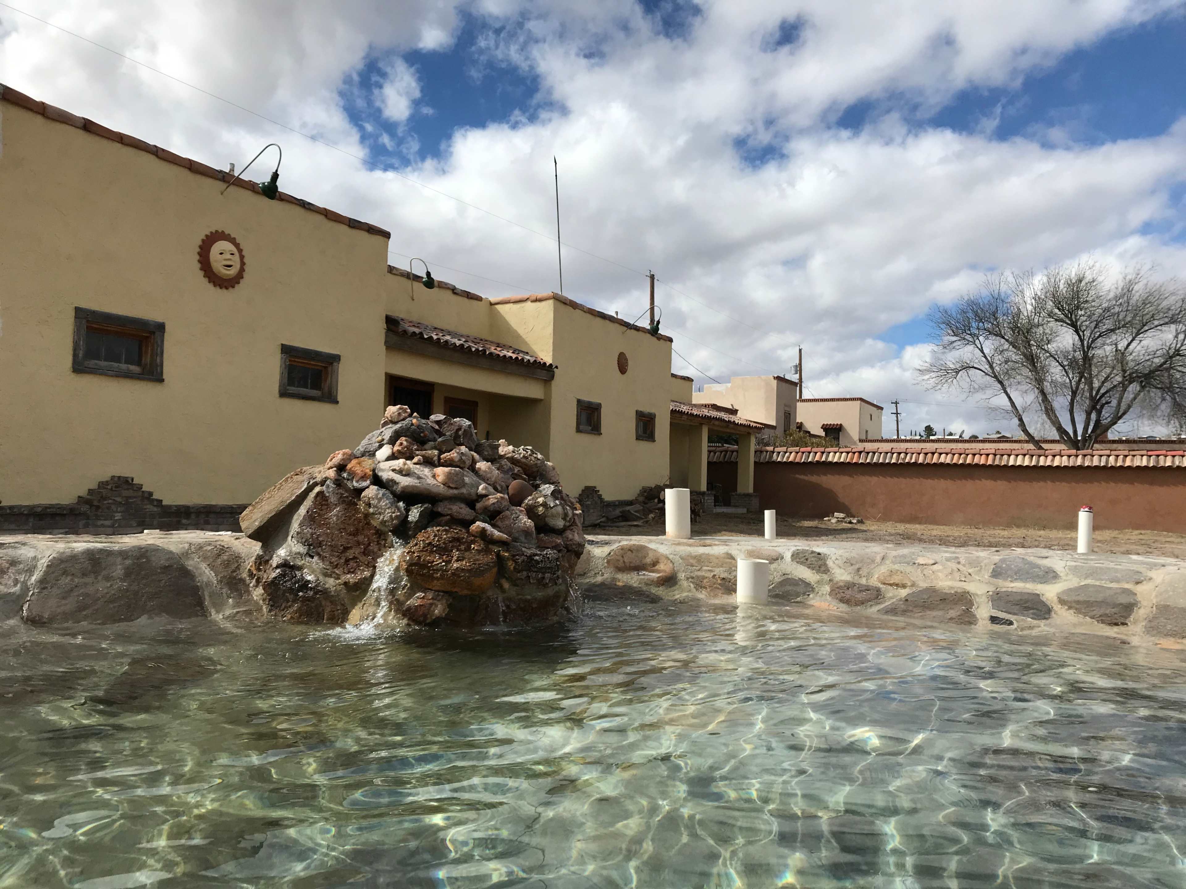 A natural hot spring surrounded by a stone border, with adobe-style buildings and a cloudy sky in the background.