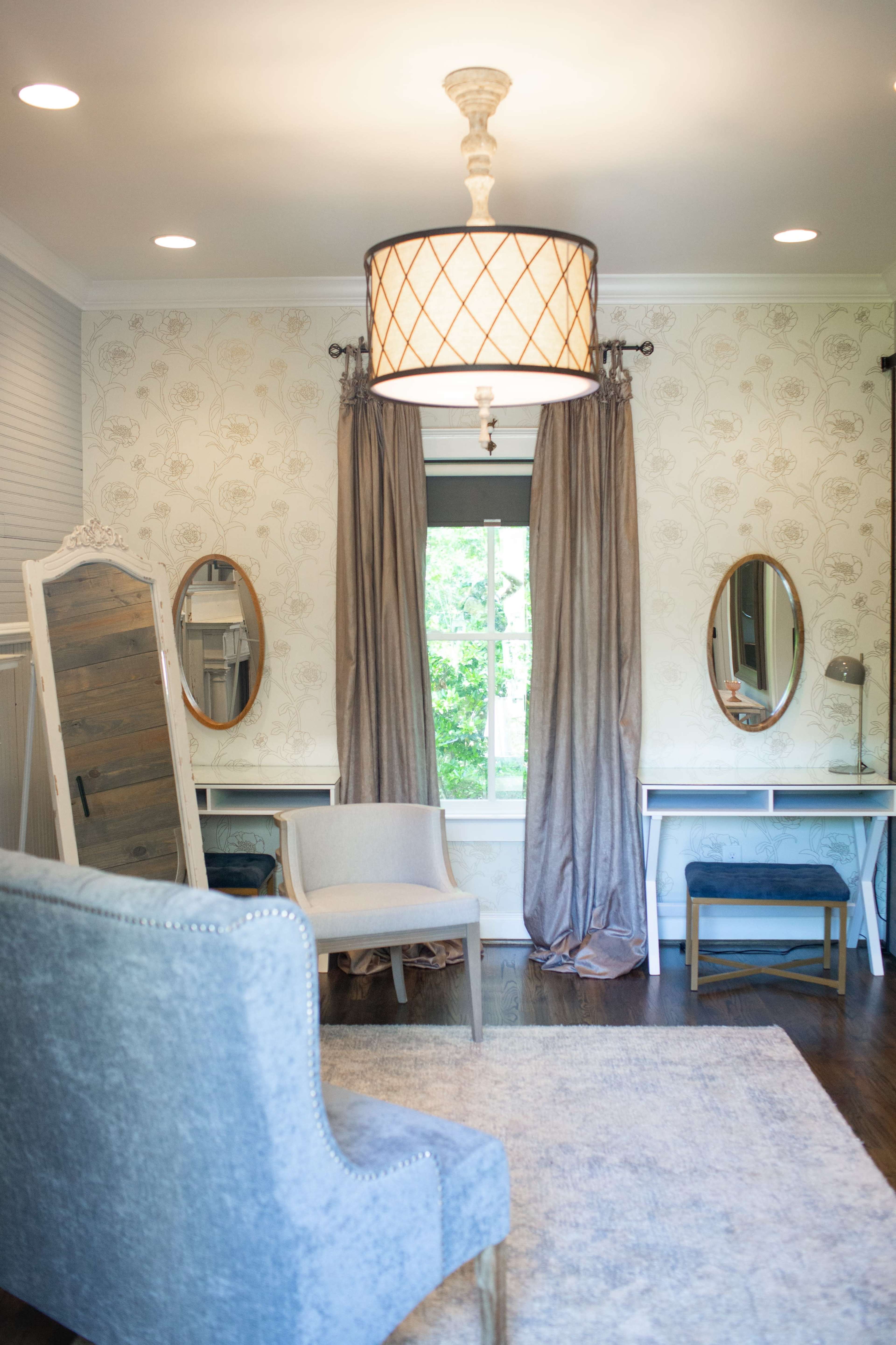A well-lit room featuring a gray armchair, a small table with a blue stool, two circular mirrors, and a large window draped with curtains.