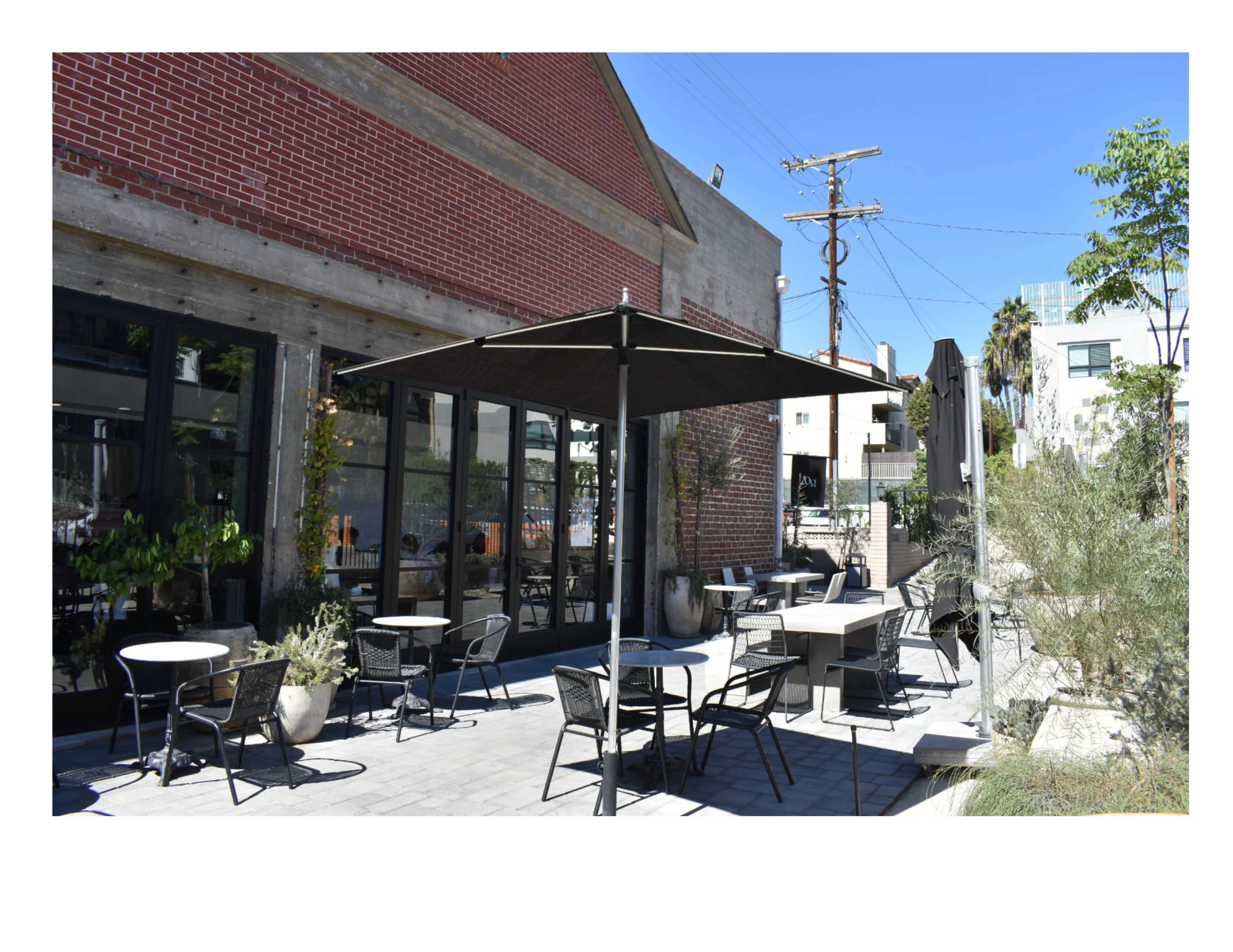 The scene shows an outdoor patio area of a restaurant with several tables and chairs, surrounded by greenery and a brick building.