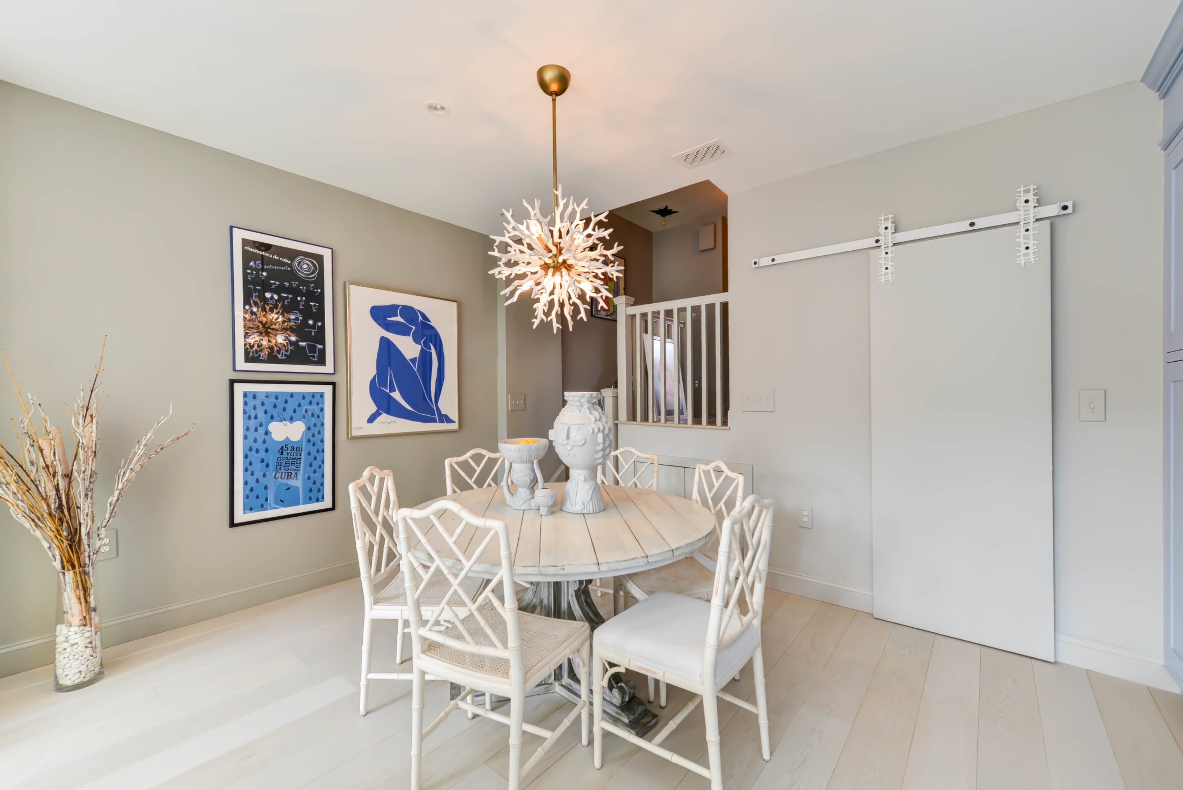 A round dining table surrounded by six chairs is set in a well-lit room featuring artwork on the walls and a sliding barn door.
