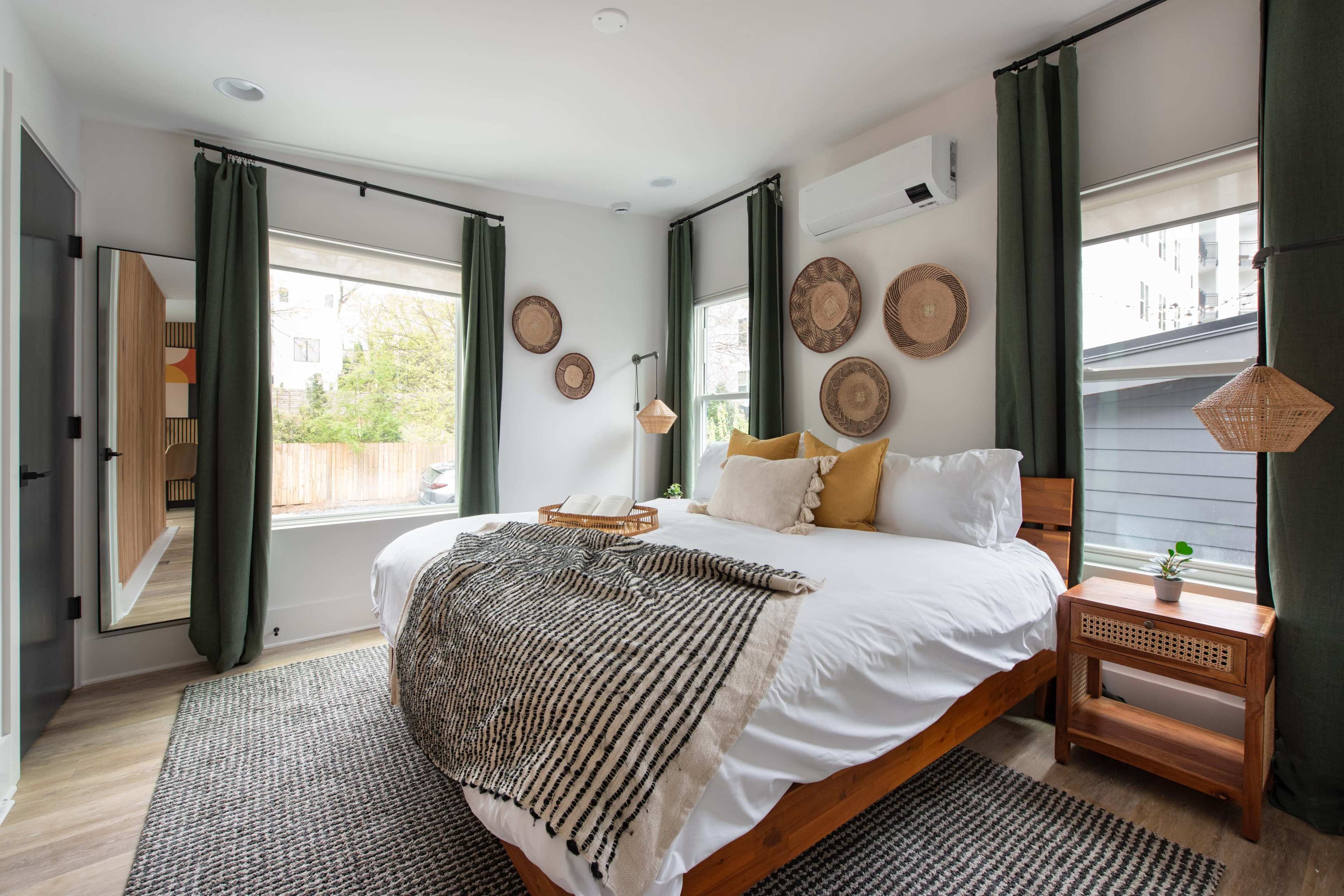 The room features a large bed with a patterned throw, two windows dressed in green curtains, and decorative wall hangings.