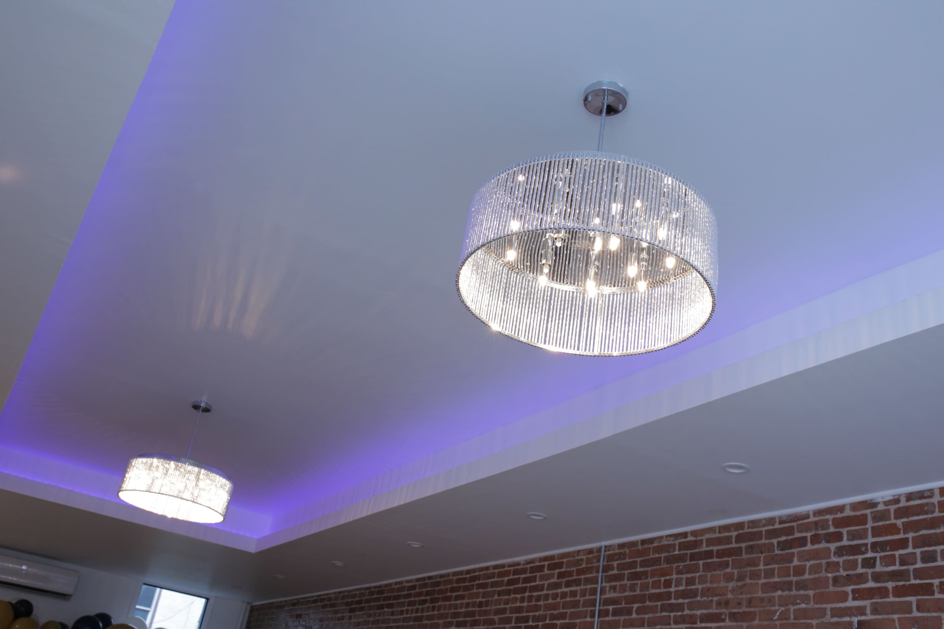 The image shows two modern chandeliers hanging from a ceiling with a light purple ambient glow above a brick wall.