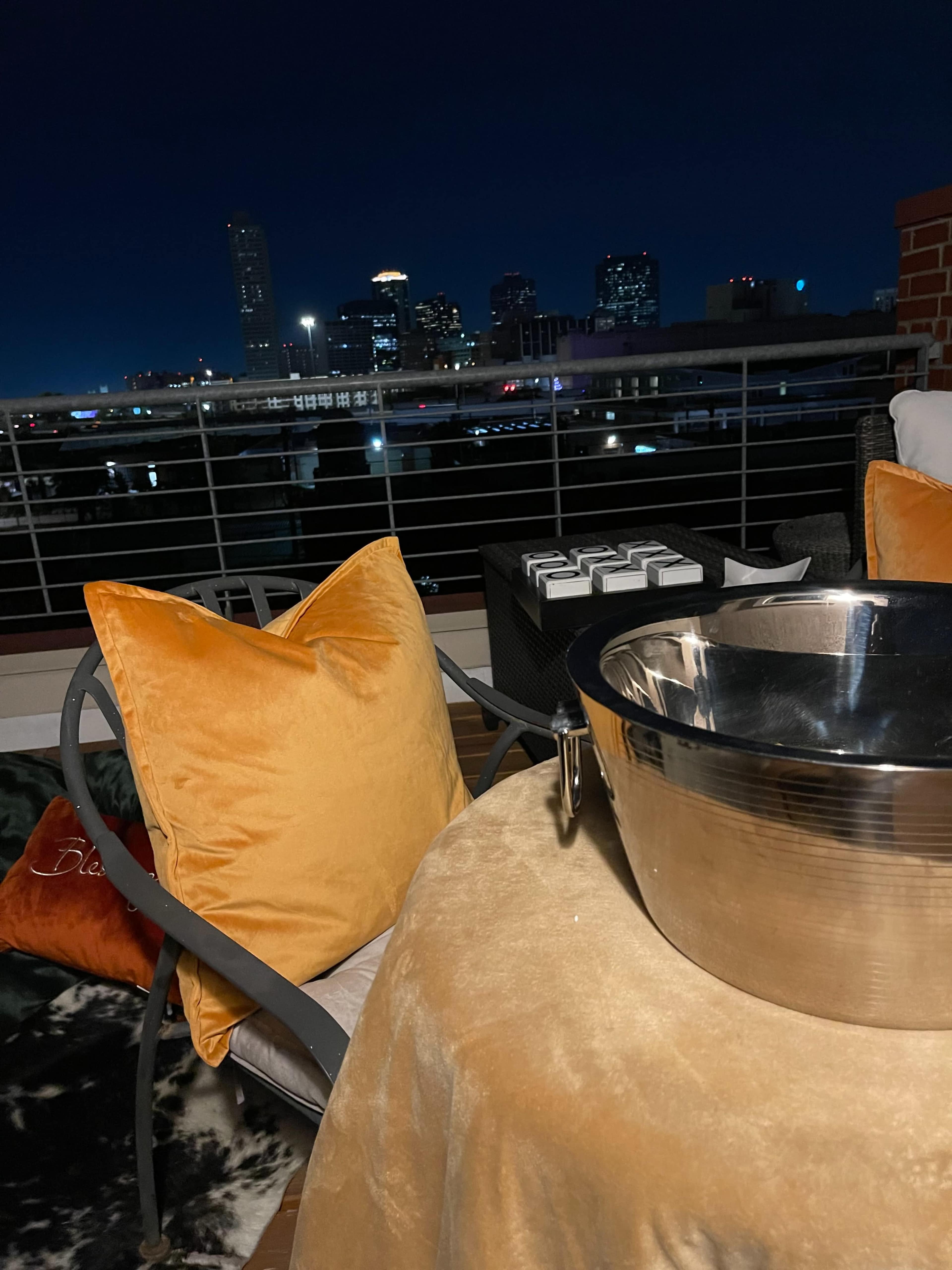 The image shows a balcony setting with a metal bowl and orange cushions overlooking a city skyline at night.