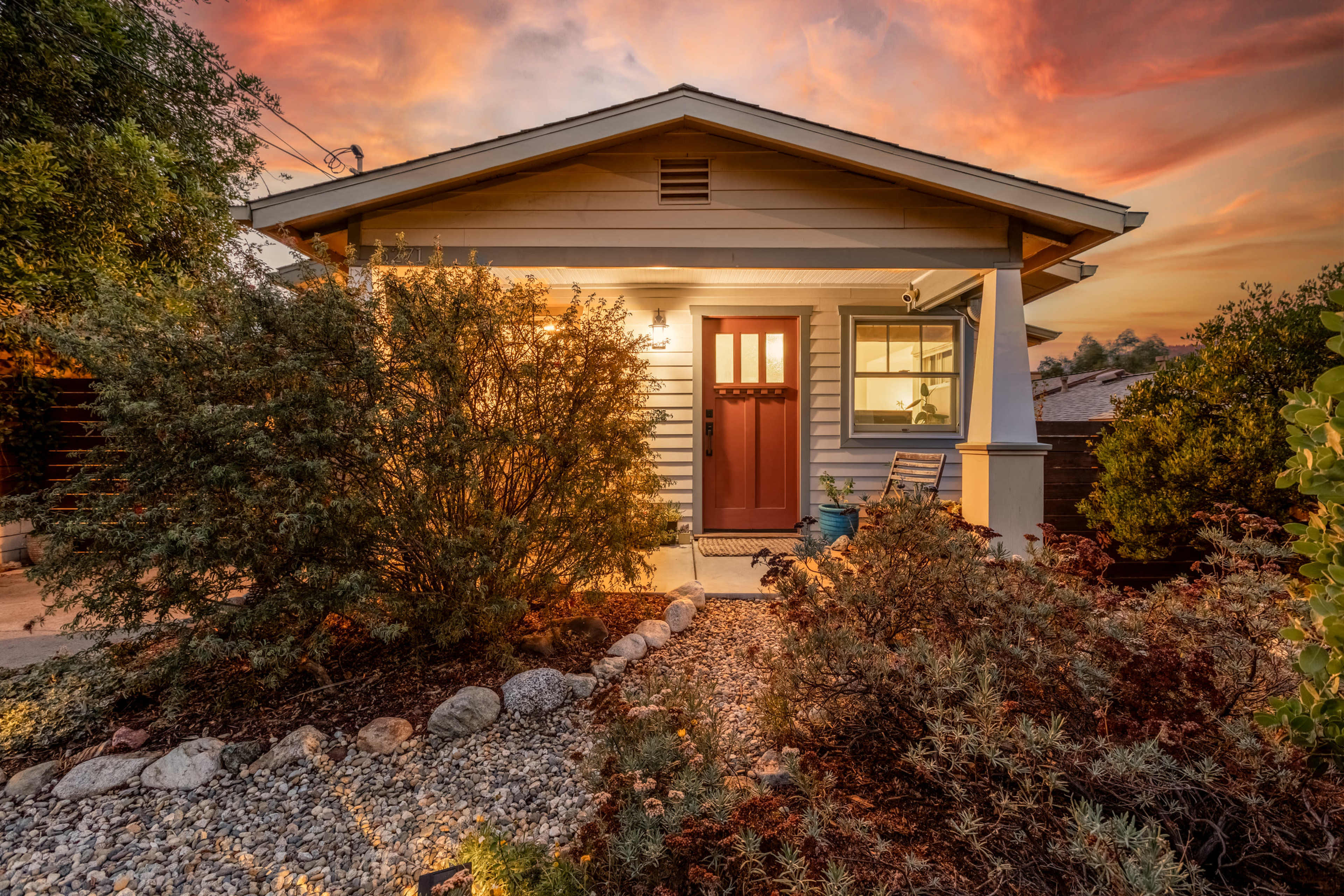 A small house with a brown front door is surrounded by landscaped gardens and rocks under a colorful sunset sky.