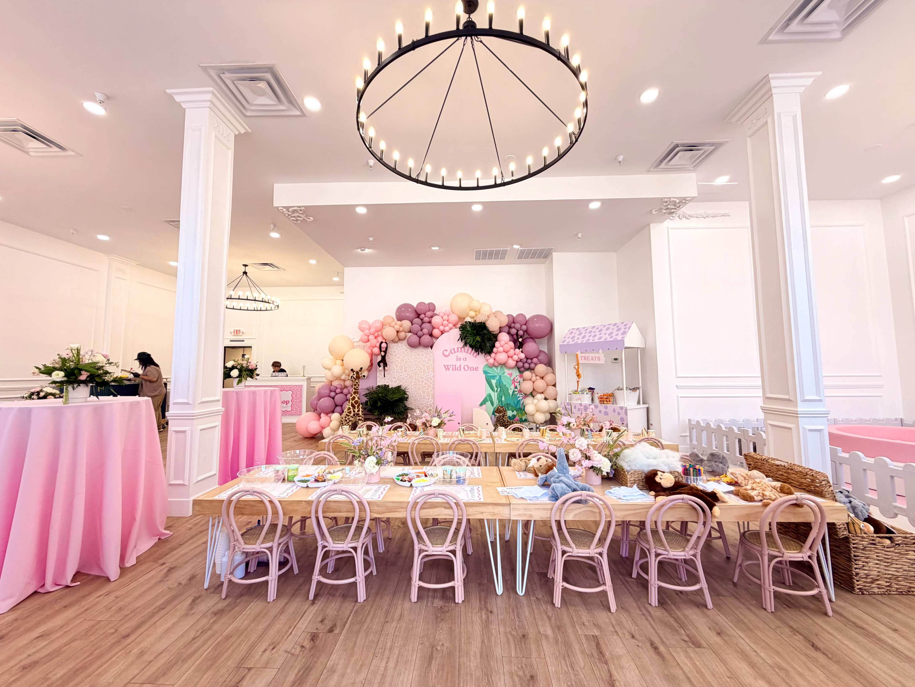 A brightly decorated event space features a long table set with pastel-colored dishes and a backdrop of balloons in shades of pink and cream.