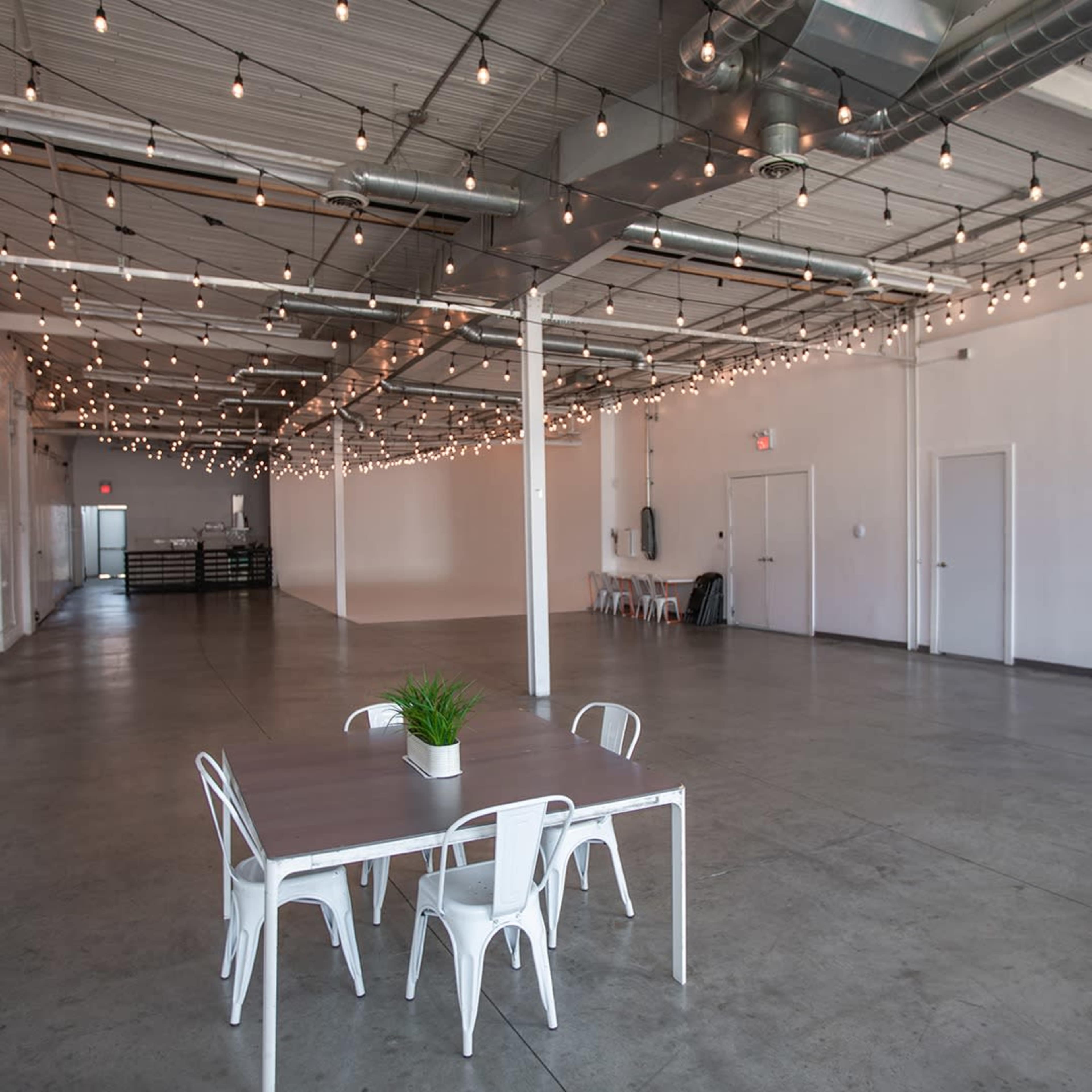 A large, empty room with polished concrete floors, string lights overhead, and a table surrounded by white chairs near the center.