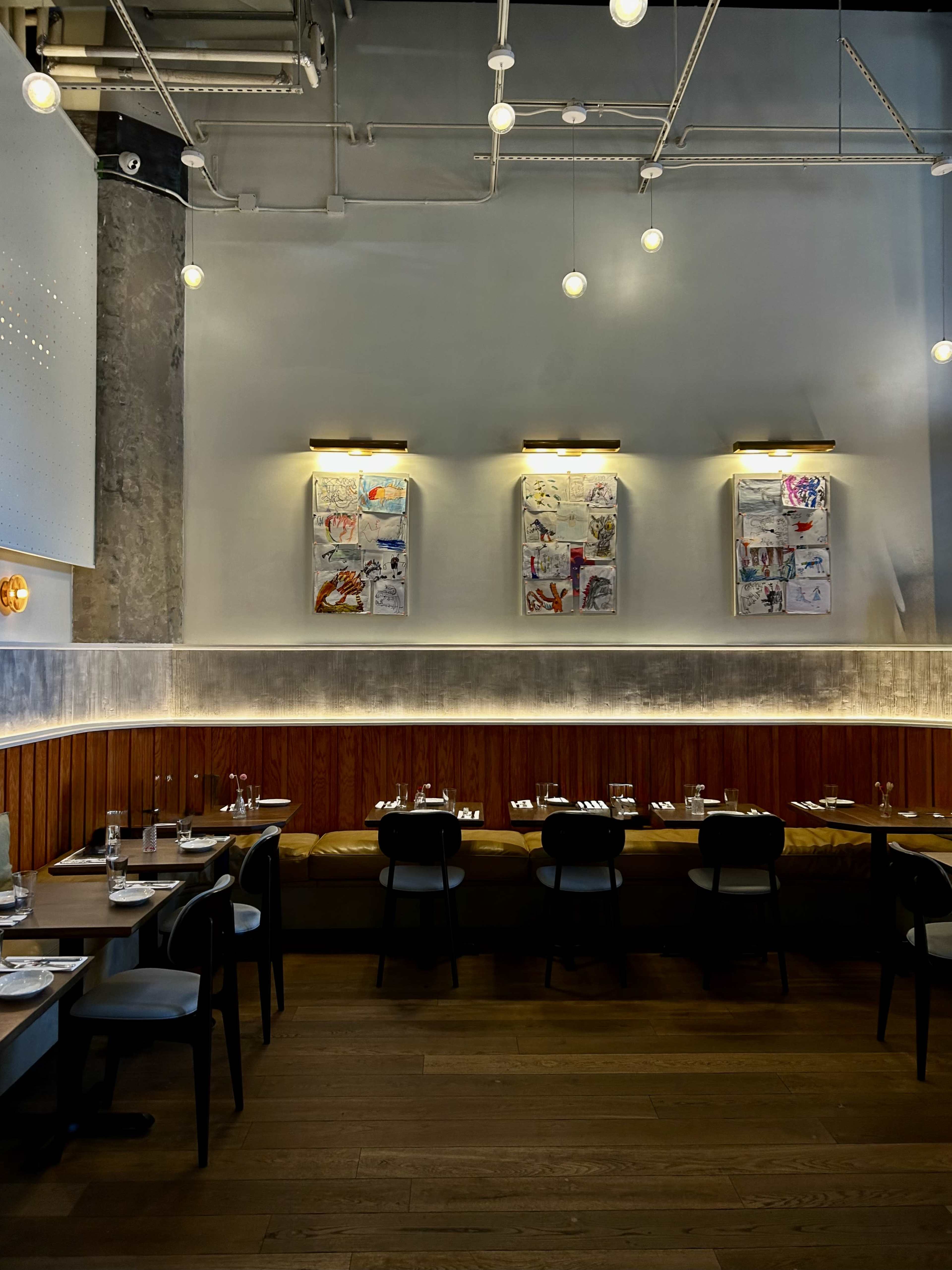 The image shows a modern restaurant interior featuring wooden tables set for dining, with a bench along the wall and framed artwork displayed above.