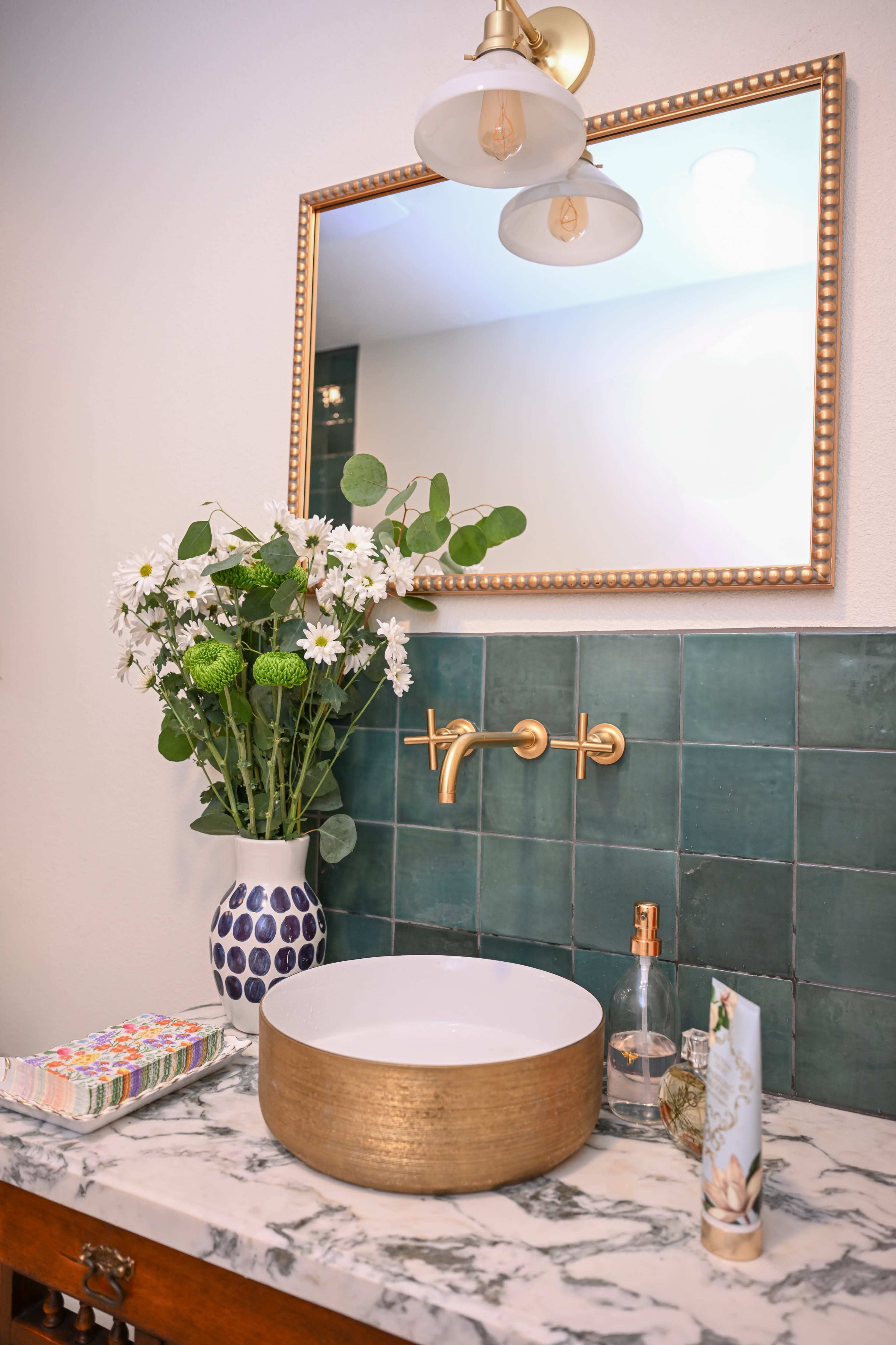 A modern bathroom features a round gold sink atop a marble countertop, with green tiled walls and a vase of flowers next to a wall-mounted faucet and mirror.
