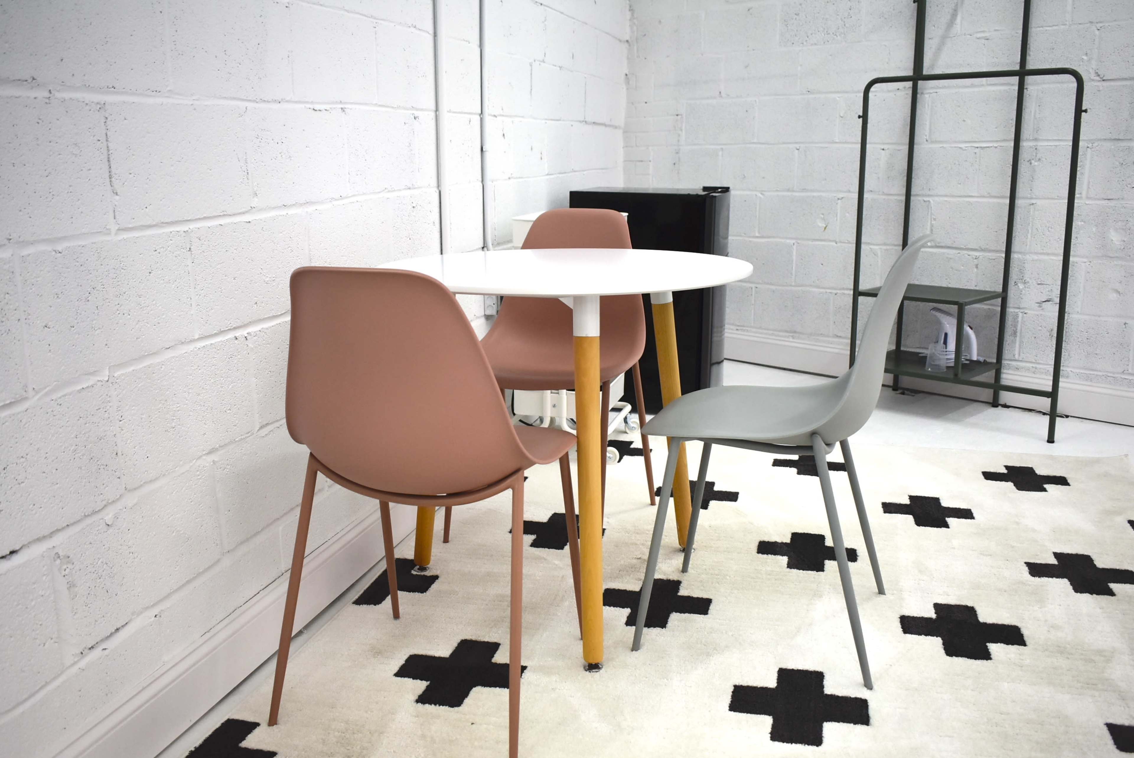 A round white table is paired with three chairs, two in muted pink and one in gray, on a patterned black and white rug in a minimalist room with a concrete wall.